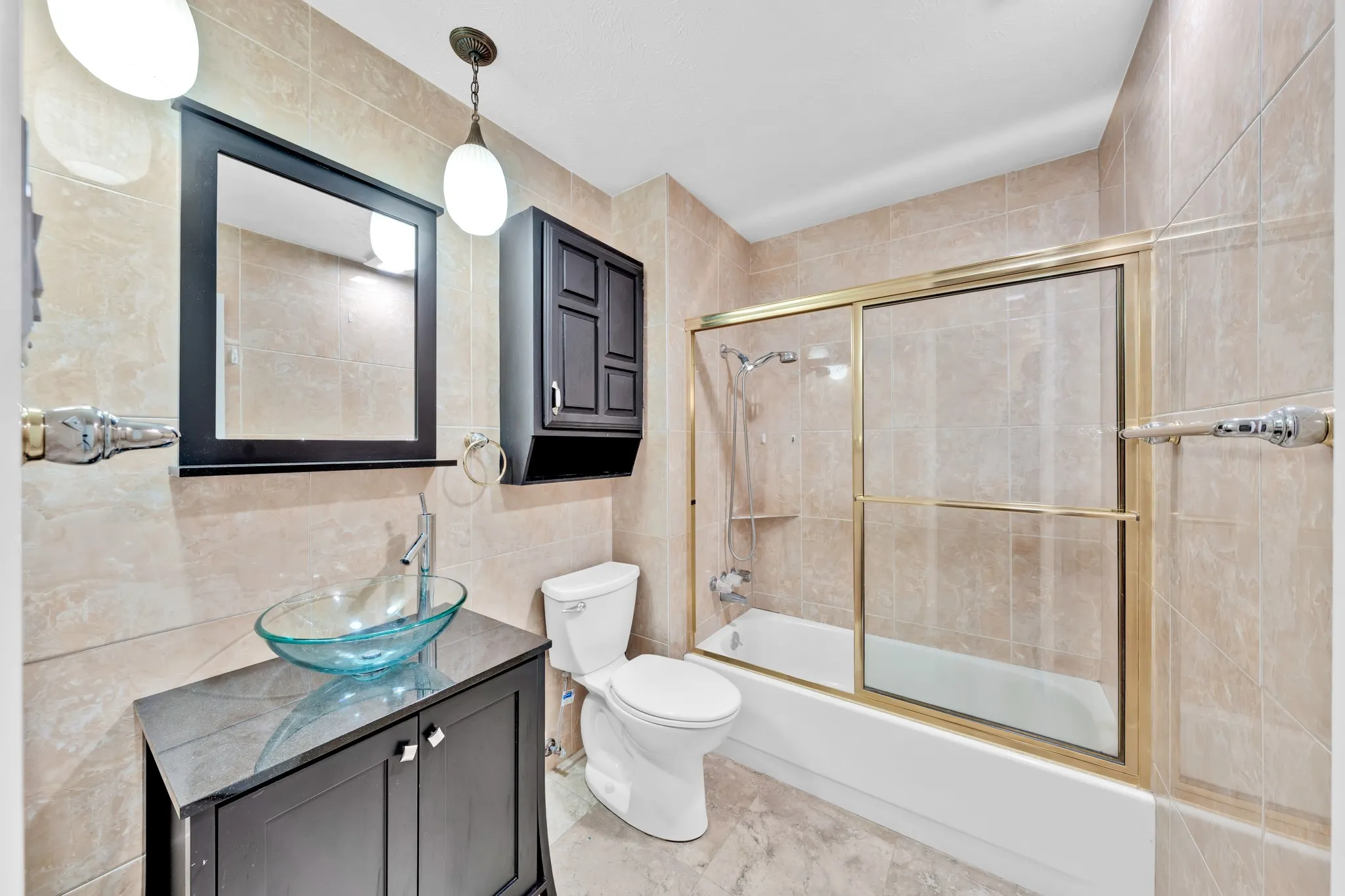Bathroom featuring full-height tiled walls, a glass vessel sink, dark wood-finish vanity, and a shower-tub combination with sliding glass doors