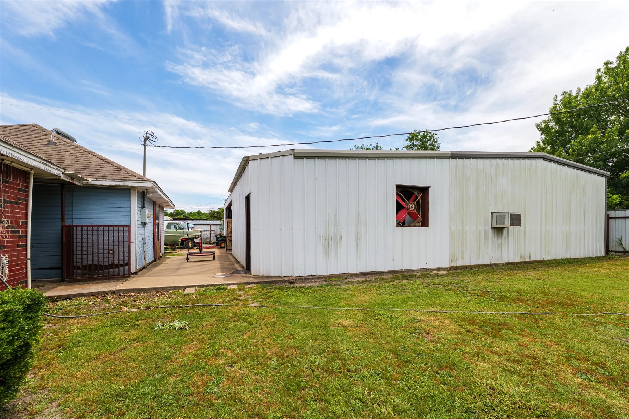 Expansive grass lawn with a concrete walkway, featuring a metal-sided outbuilding with an exterior fan and an additional structure with brick and blue siding
