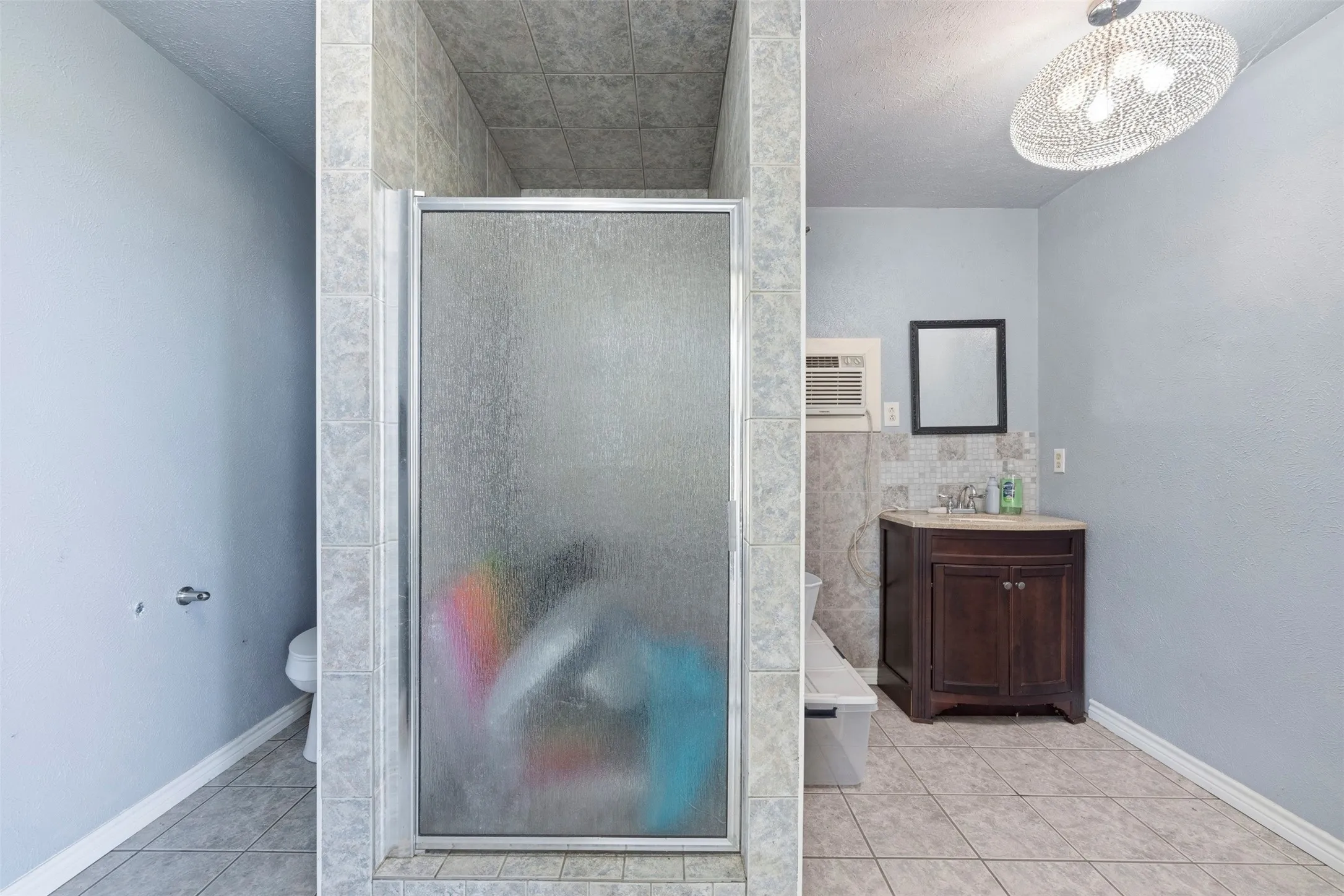 Frosted glass shower enclosure, wood-finish vanity with countertop, tiled flooring, and decorative overhead lighting