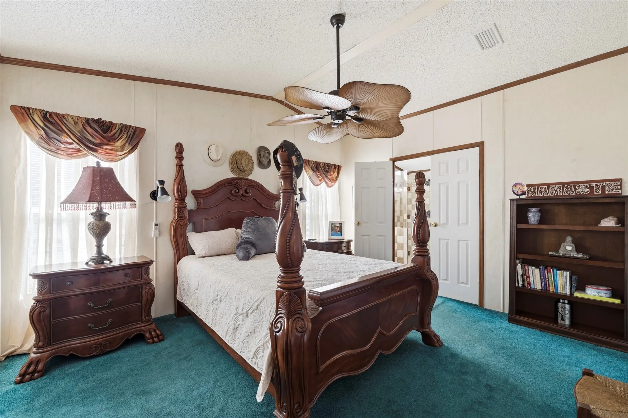 Ceiling fan with unique blade design, white paneled walls with wood trim, light-colored window treatments, and teal carpet flooring