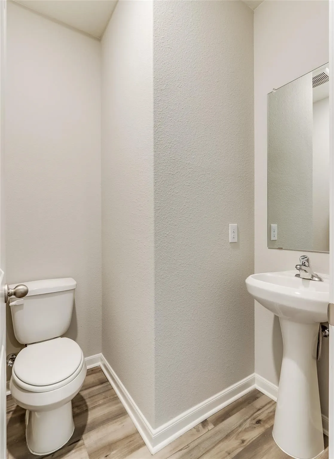 Pedestal sink with chrome faucet, wall-mounted mirror, white toilet, and wood-finish flooring