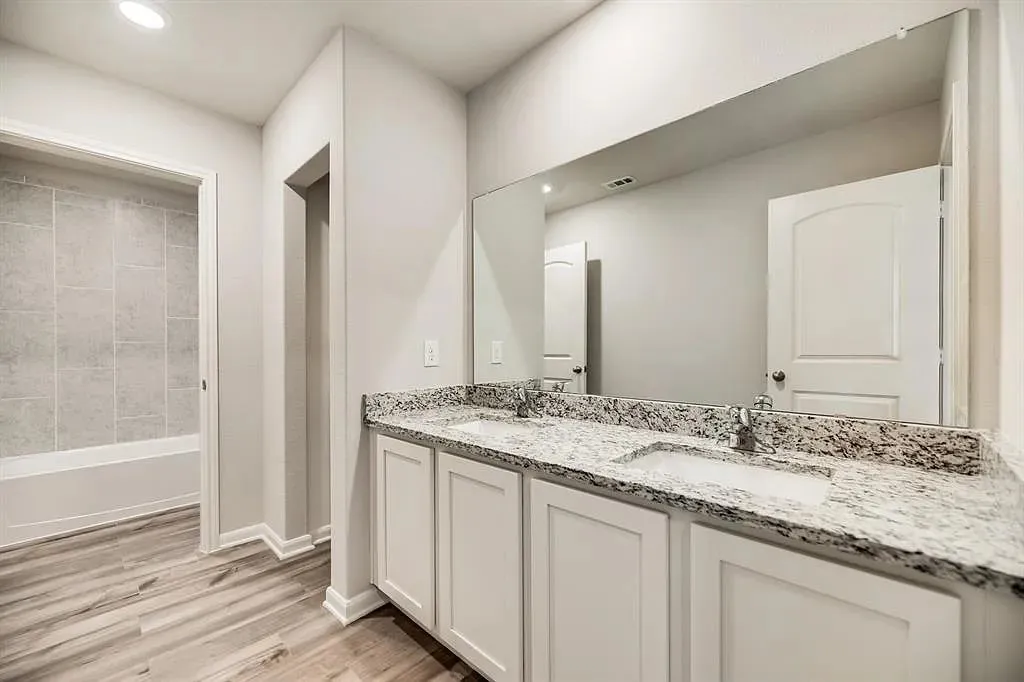 Dual vanity with integrated sinks, granite countertop, white cabinetry, wood-finish flooring, and a tiled shower/tub combination