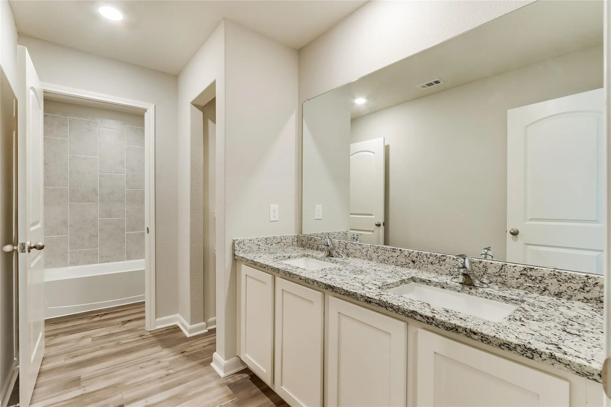 Dual vanity with granite countertop, integrated sinks, chrome fixtures, wood-finish flooring, and a full-width mirror