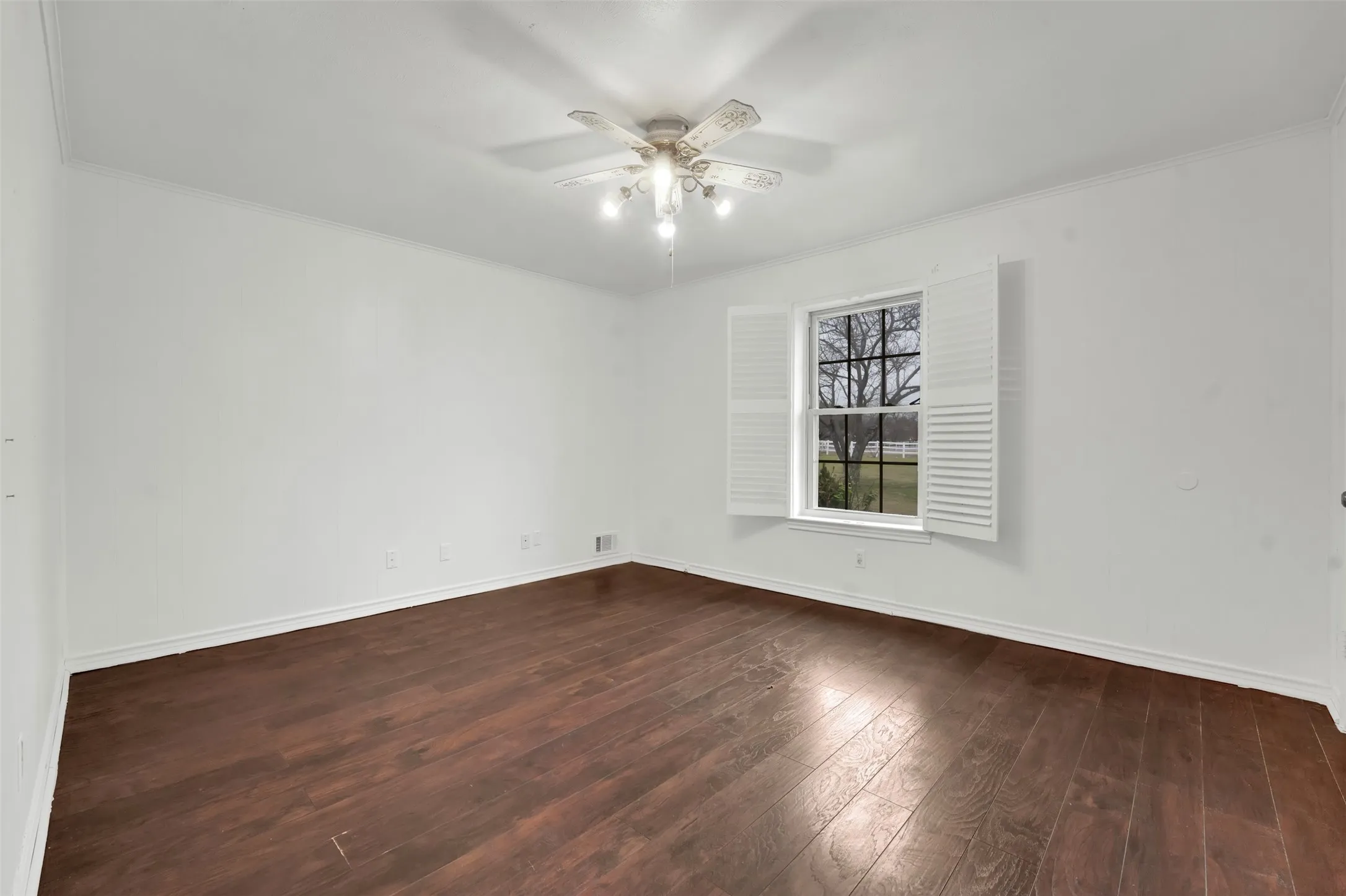 Spacious room featuring rich wood-finish flooring, white walls, and abundant natural light from a large window with plantation shutters