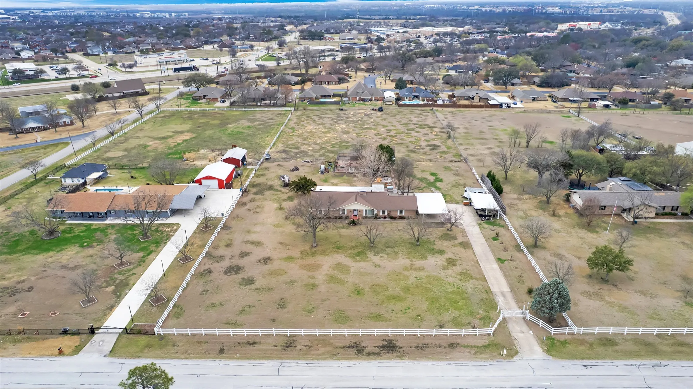 Expansive rural acreage featuring multiple outbuildings, including a red metal-roof structure and brown residence, set on fenced pastureland with mature trees