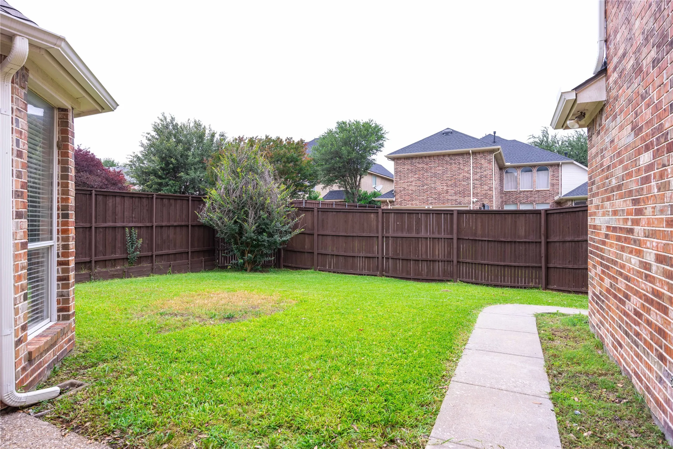 Fenced backyard with a manicured lawn, concrete pathway, and mature landscaping