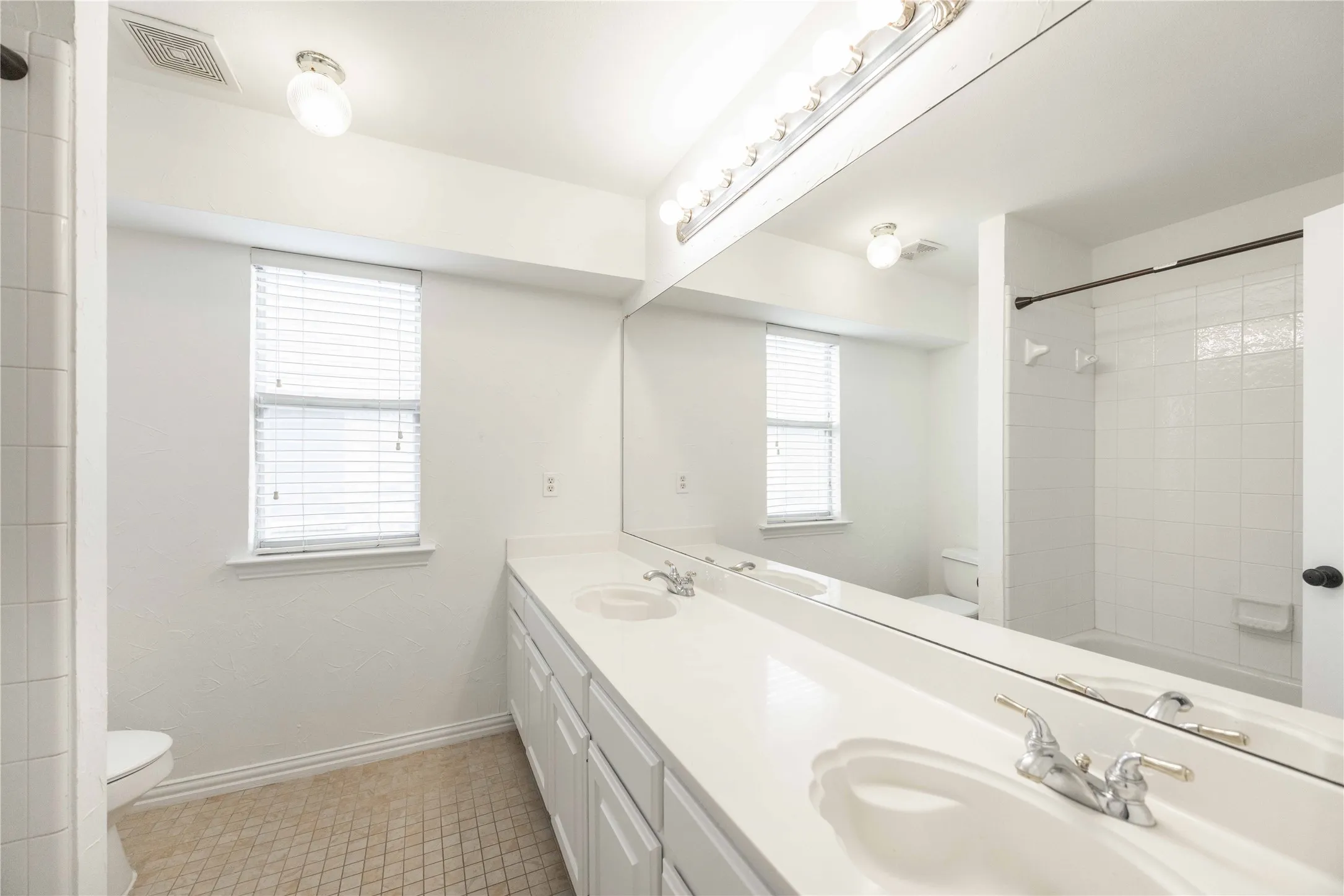 Dual vanity with integrated sinks, expansive mirror, a window with blinds, and white tile tub/shower combination