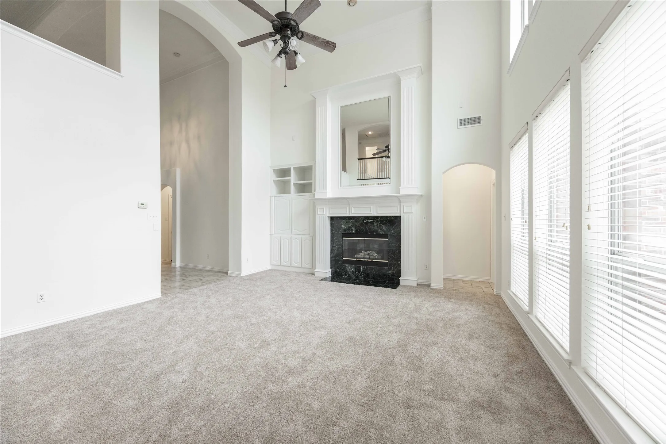 Spacious living area featuring high ceilings, a fireplace with a dark stone surround, tall windows with blinds, a ceiling fan, and light-toned carpeting
