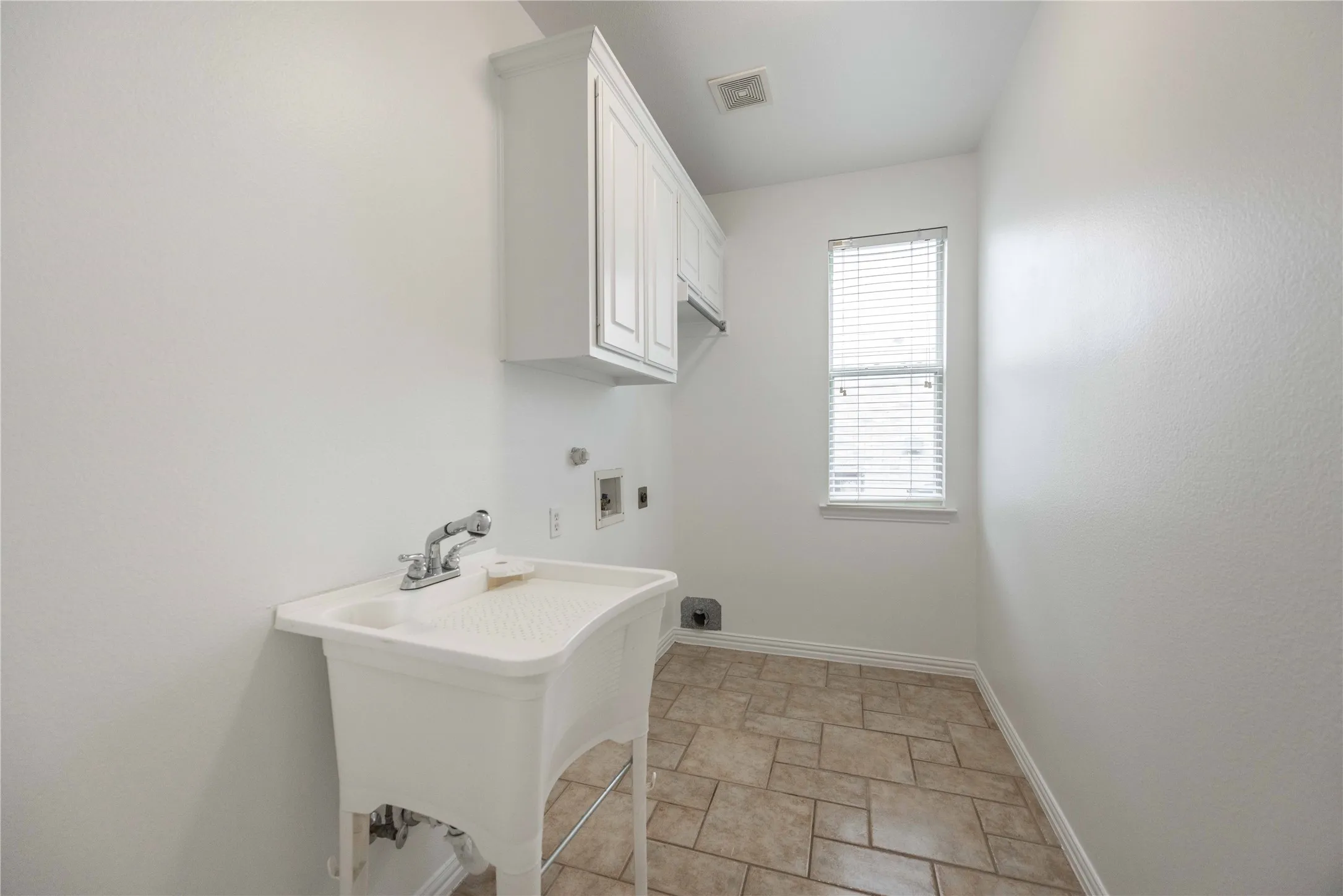 Utility sink, upper cabinetry, window with blinds, and tile flooring