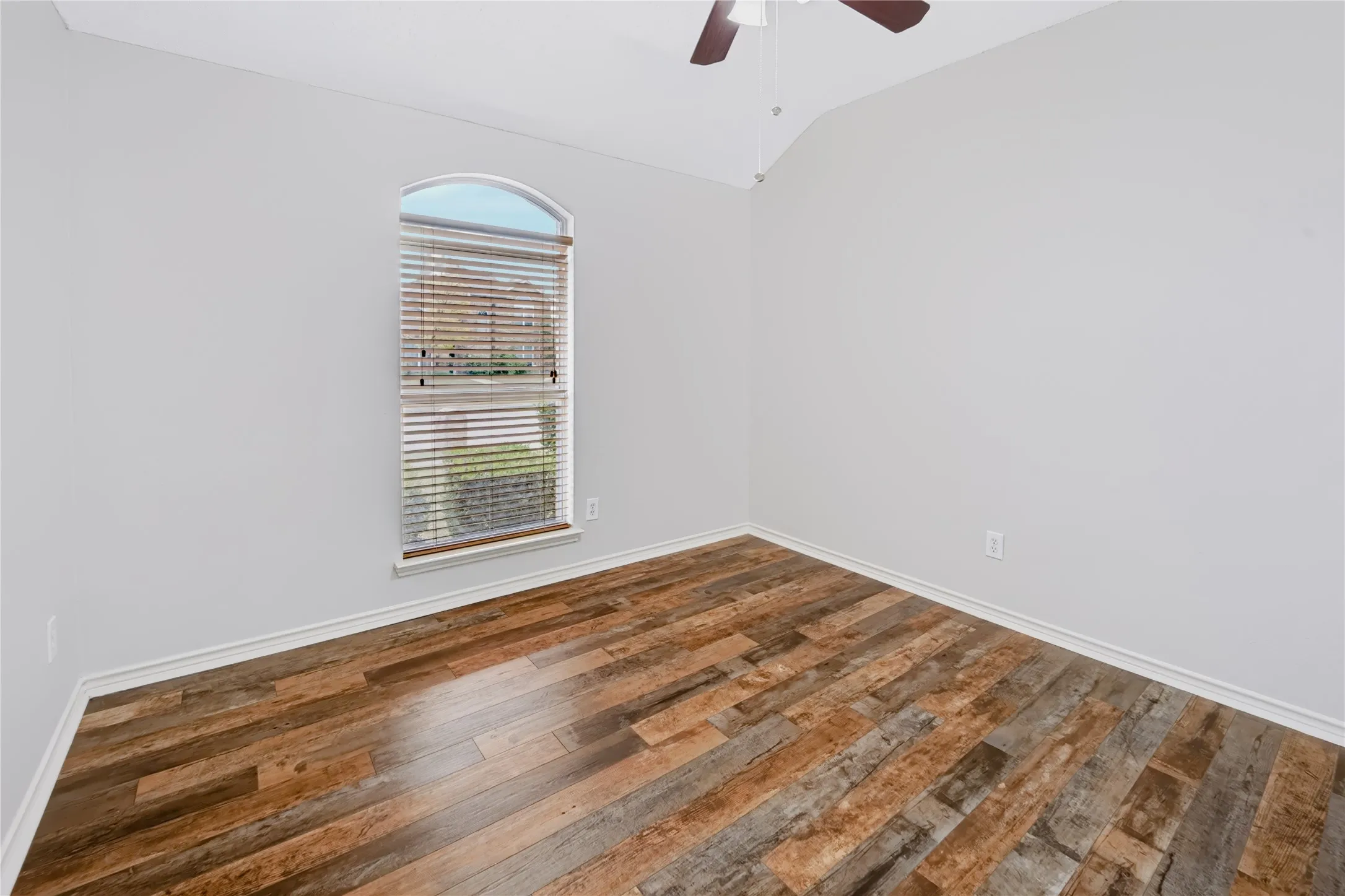 Vaulted ceiling, wood-finish flooring, and arched window with integrated blinds, complemented by a ceiling fan