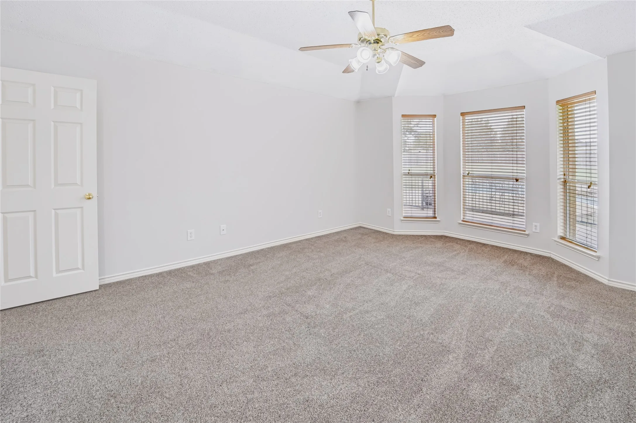 Vaulted ceiling with a central multi-blade ceiling fan, wall-to-wall carpeting, and a three-window bay configuration featuring horizontal blinds