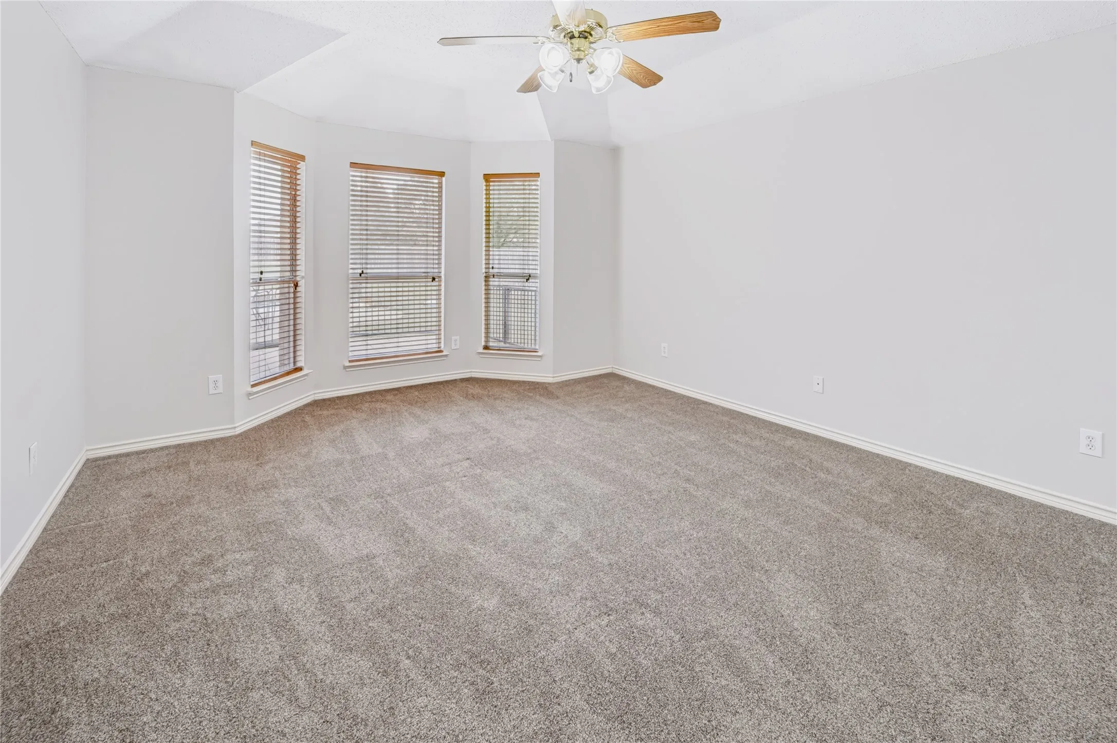 Room featuring high tray ceilings, neutral carpeting, three large windows with horizontal blinds, and a central ceiling fan with integrated lighting