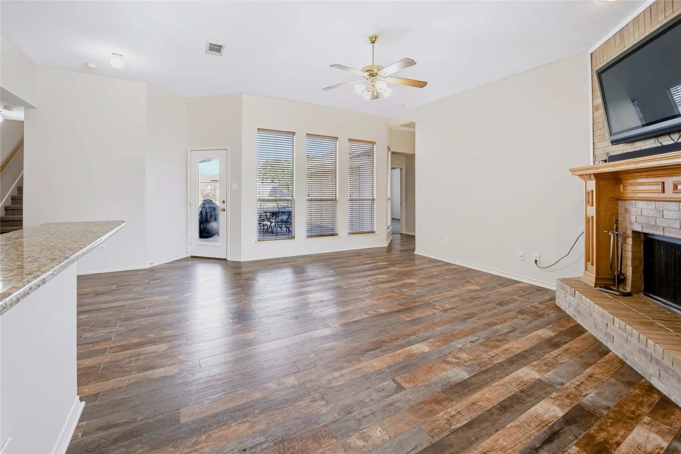 Open-concept interior featuring wood-finish tile flooring, a floor-to-ceiling brick fireplace with a built-in wooden mantel, triple vertical windows, and a raised breakfast bar with a stone-finish countertop