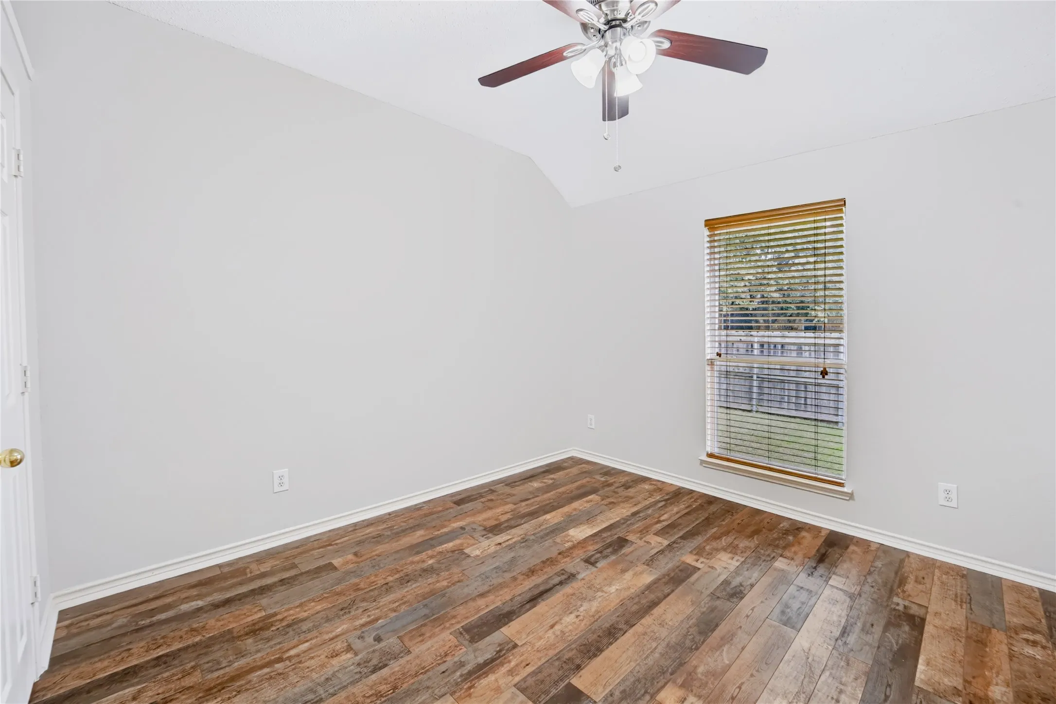 Vaulted ceiling featuring a central lighted ceiling fan, wood-finish flooring, and a single window with horizontal blinds