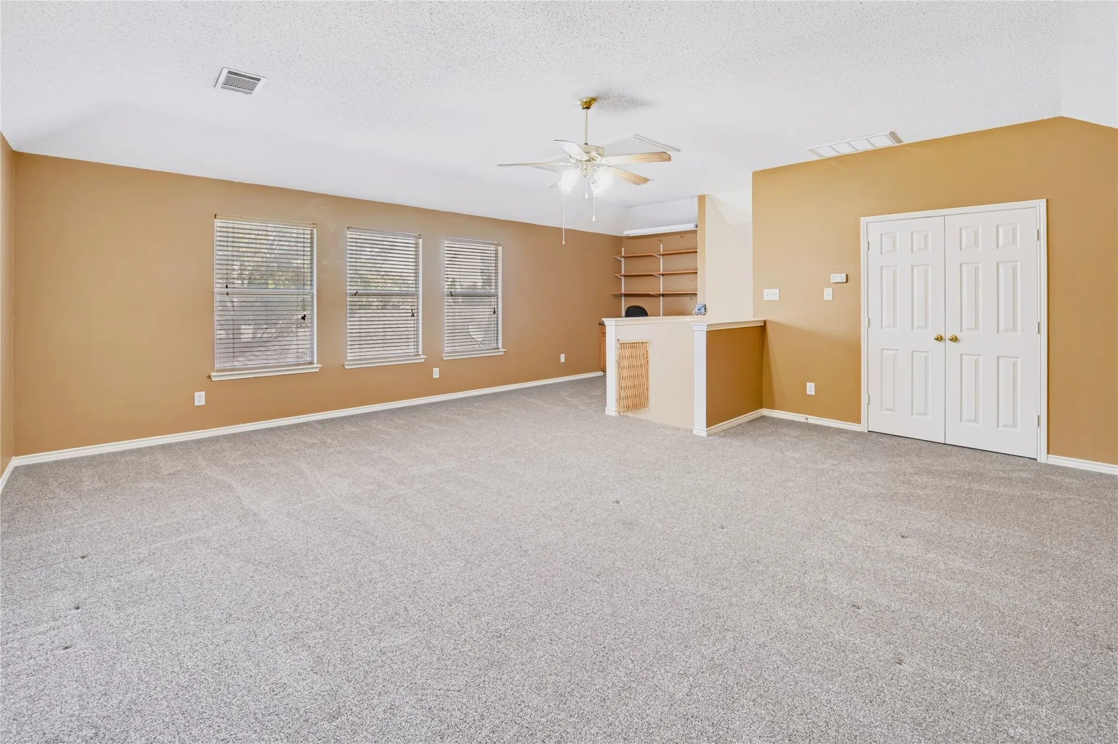 Spacious room featuring a vaulted ceiling, three windows with horizontal blinds, built-in wooden shelving, and double-door entry