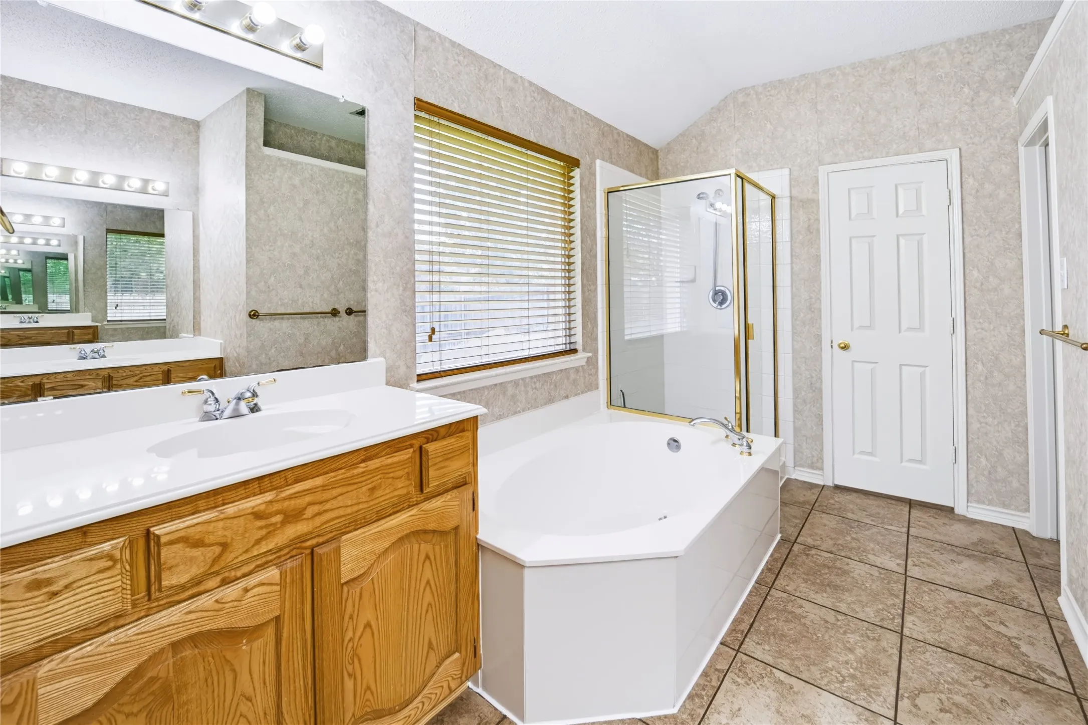 Bathroom featuring a wooden vanity, integrated countertop with a basin sink, corner soaking tub, glass-enclosed shower, and tiled flooring