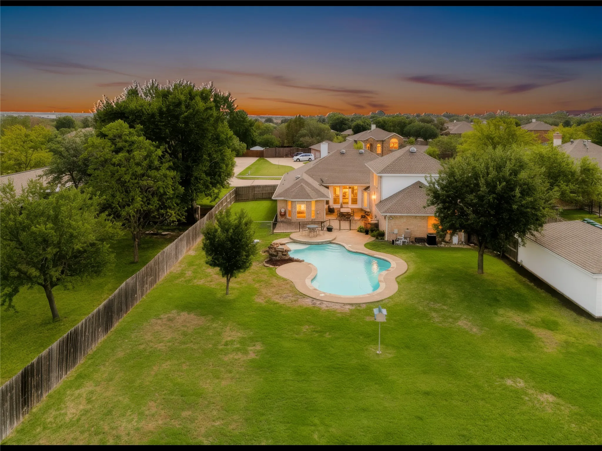 Expansive backyard featuring a free-form swimming pool, rock waterfall feature, concrete patio, mature trees, and wood privacy fencing