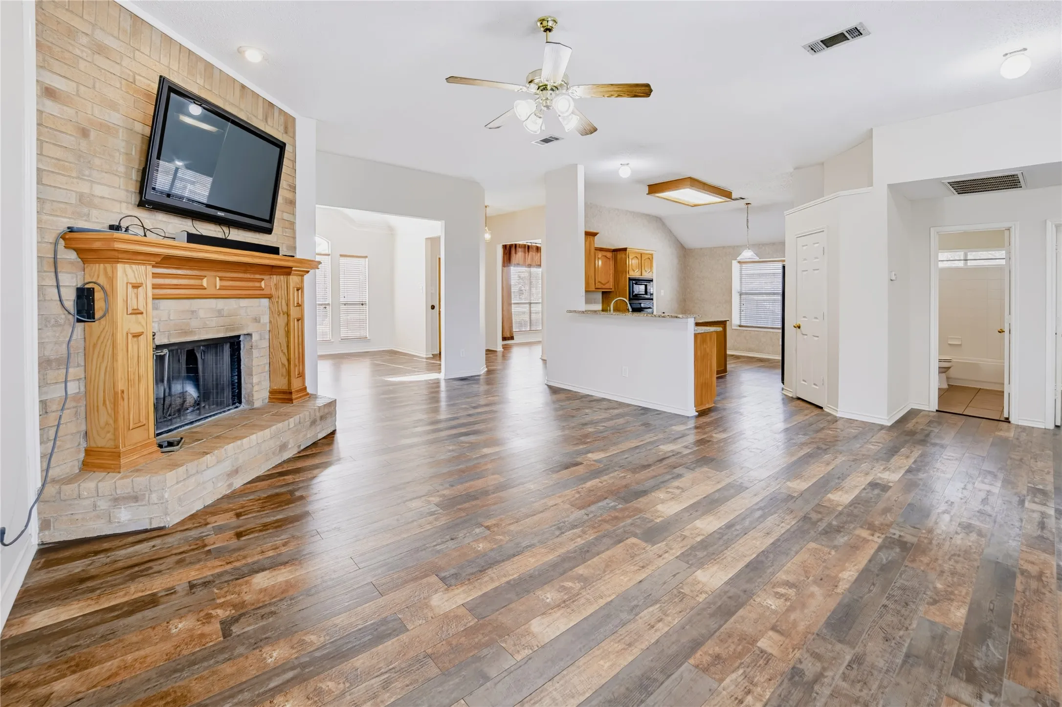 Brick fireplace with a wood mantel, wood-finish flooring, ceiling fan, and built-in kitchen cabinetry