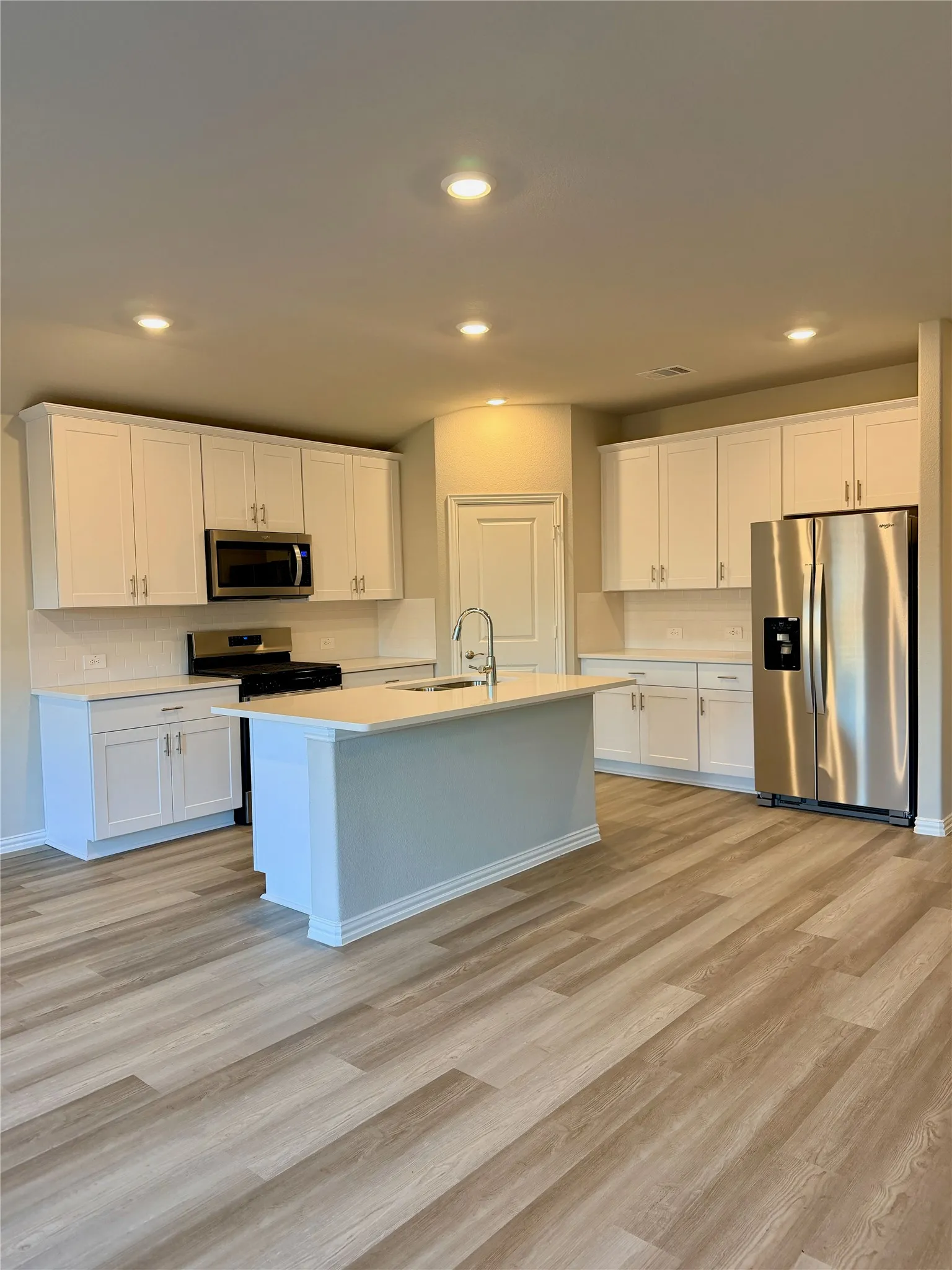 Kitchen featuring white cabinetry, light-toned countertops, stainless steel appliances, a center island with a sink, and wood-finish flooring