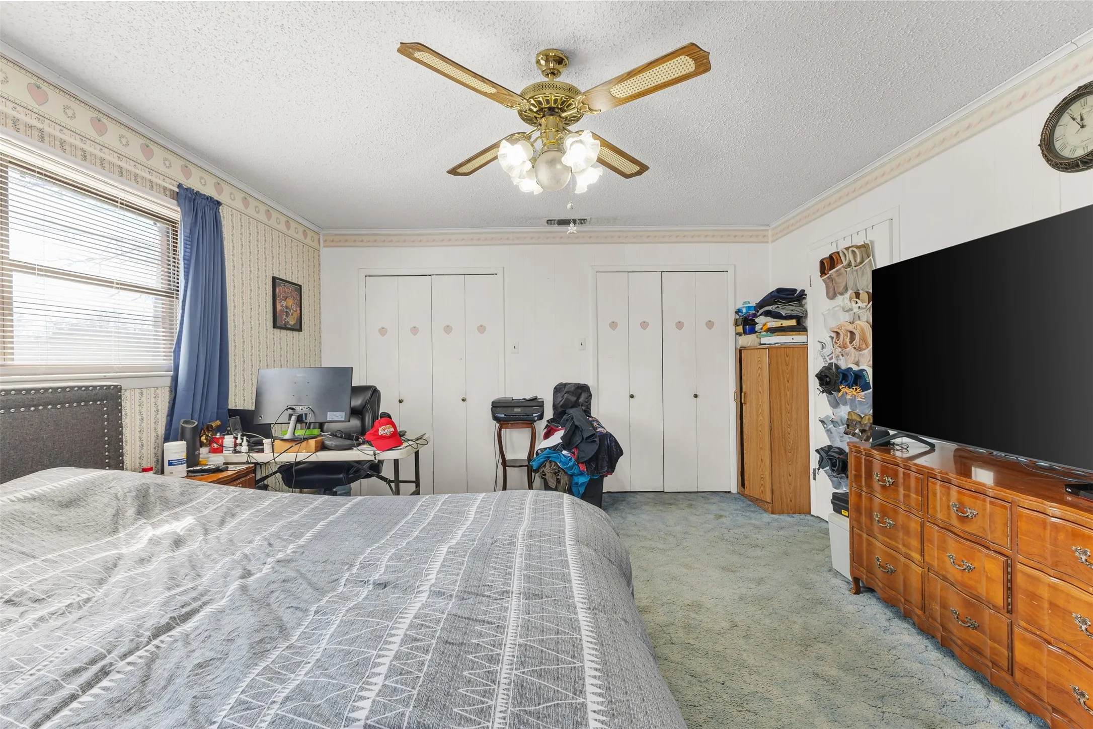 Ceiling fan with light fixture, window with blinds, dual bi-fold closet doors, wall-mounted clock, and built-in wooden shelving unit