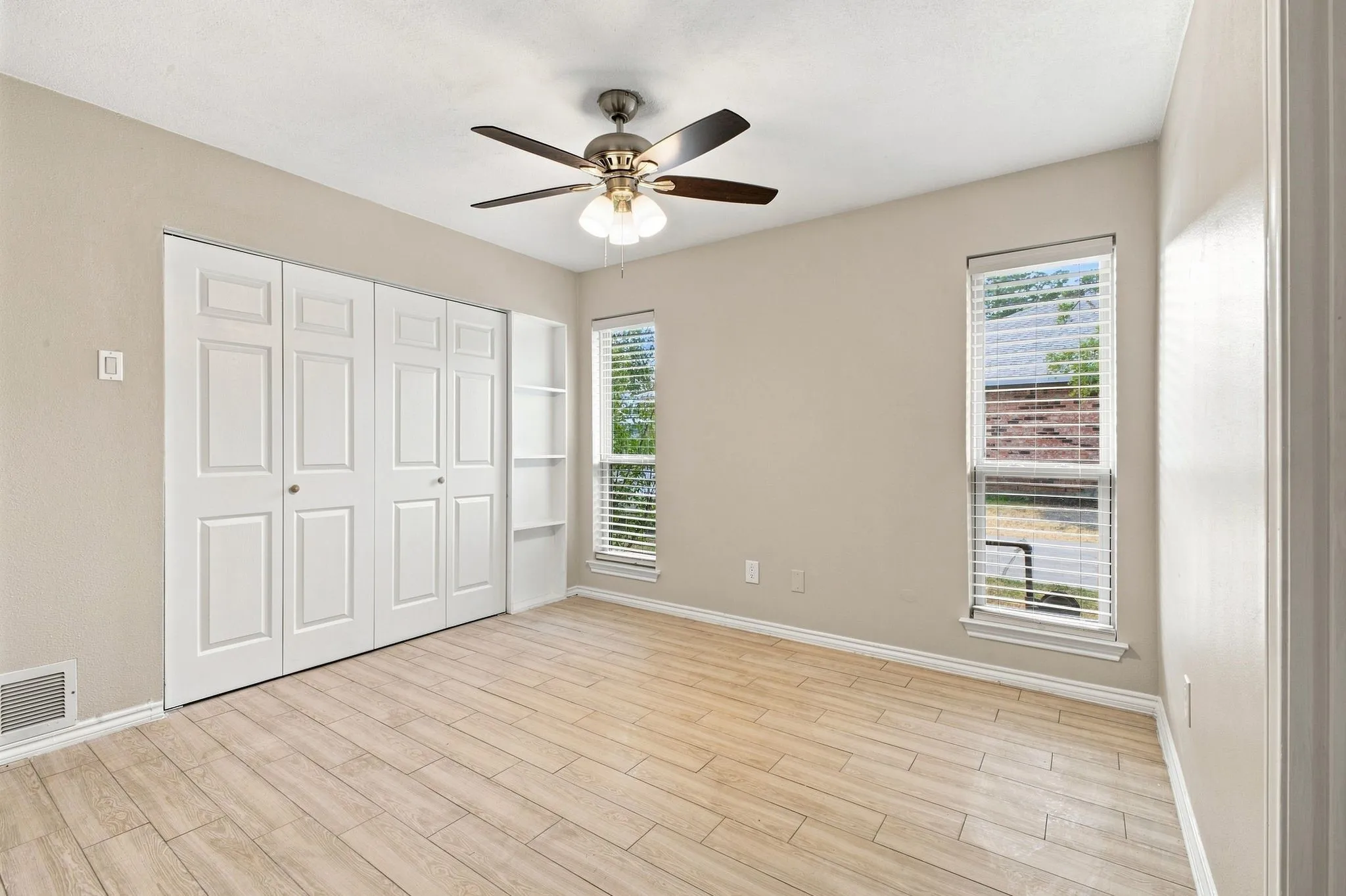 Neutral wall tones, wood-finish flooring, two large windows with blinds, a ceiling fan with integrated lighting, and built-in closet storage with bi-fold doors