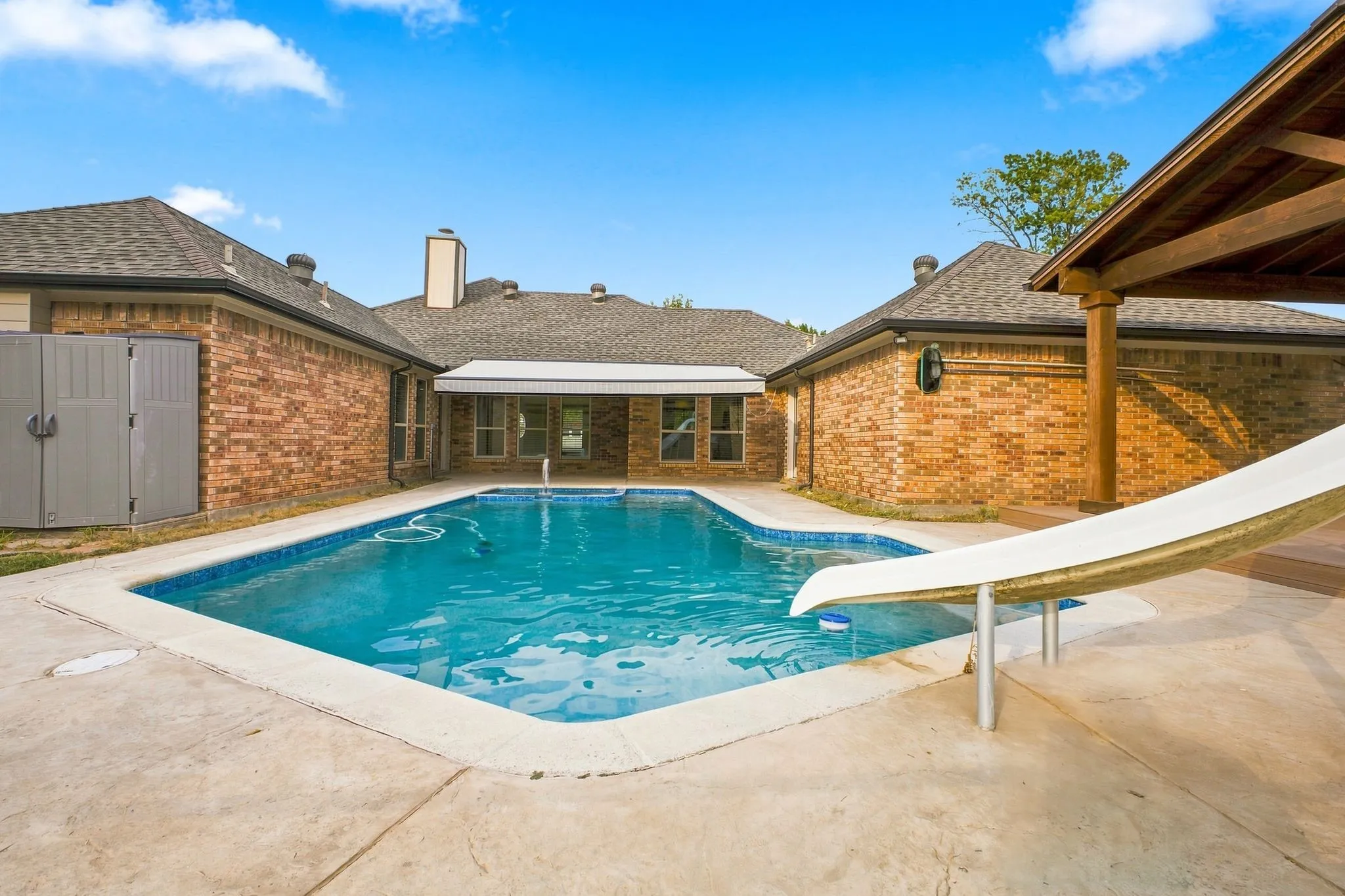 In-ground swimming pool with a slide, brick exterior, covered patio, and concrete decking