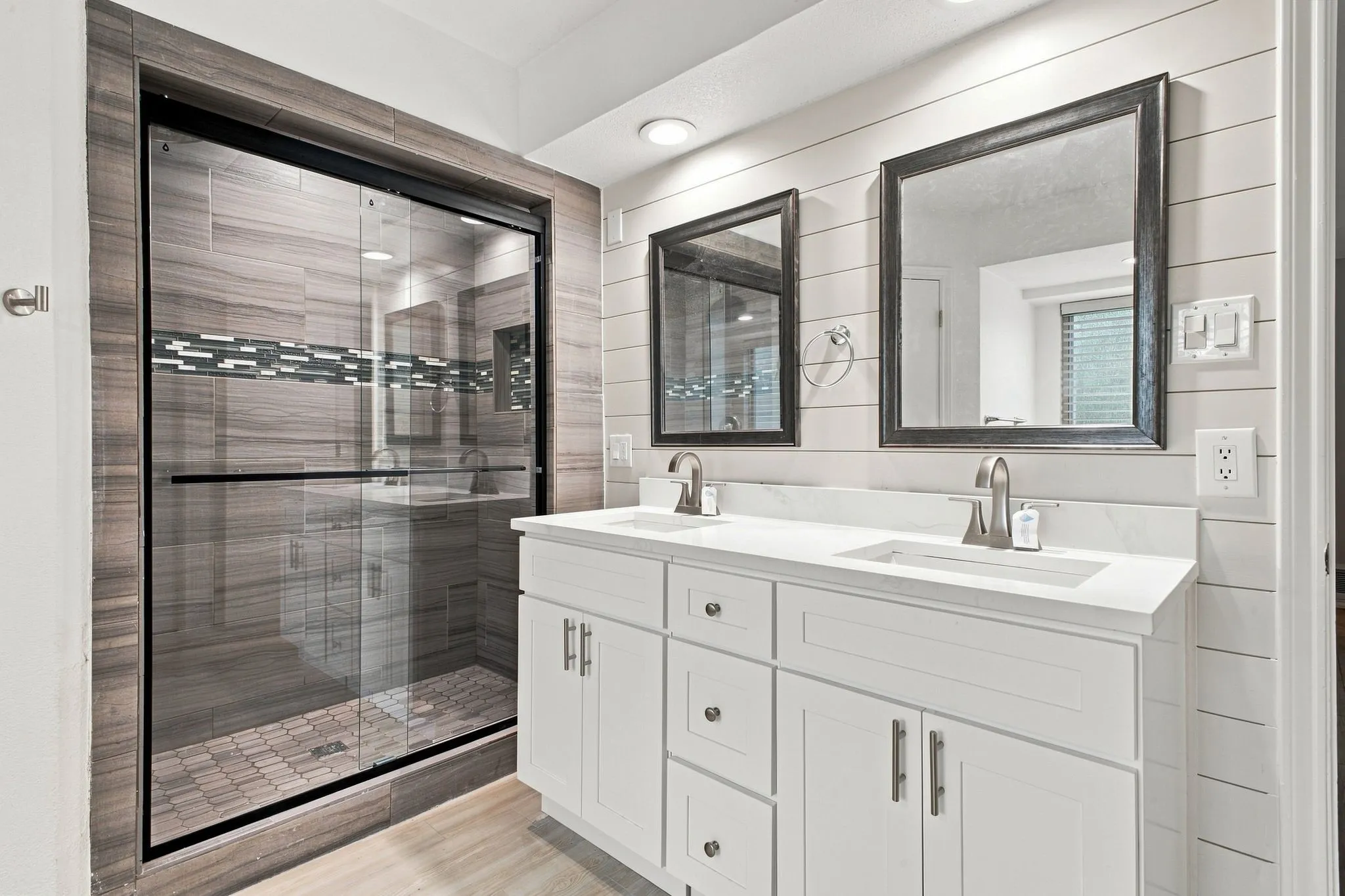 Dual vanity with white cabinetry and integrated sinks, glass-enclosed shower with tile surround, shiplap wall accents, and wood-finish flooring
