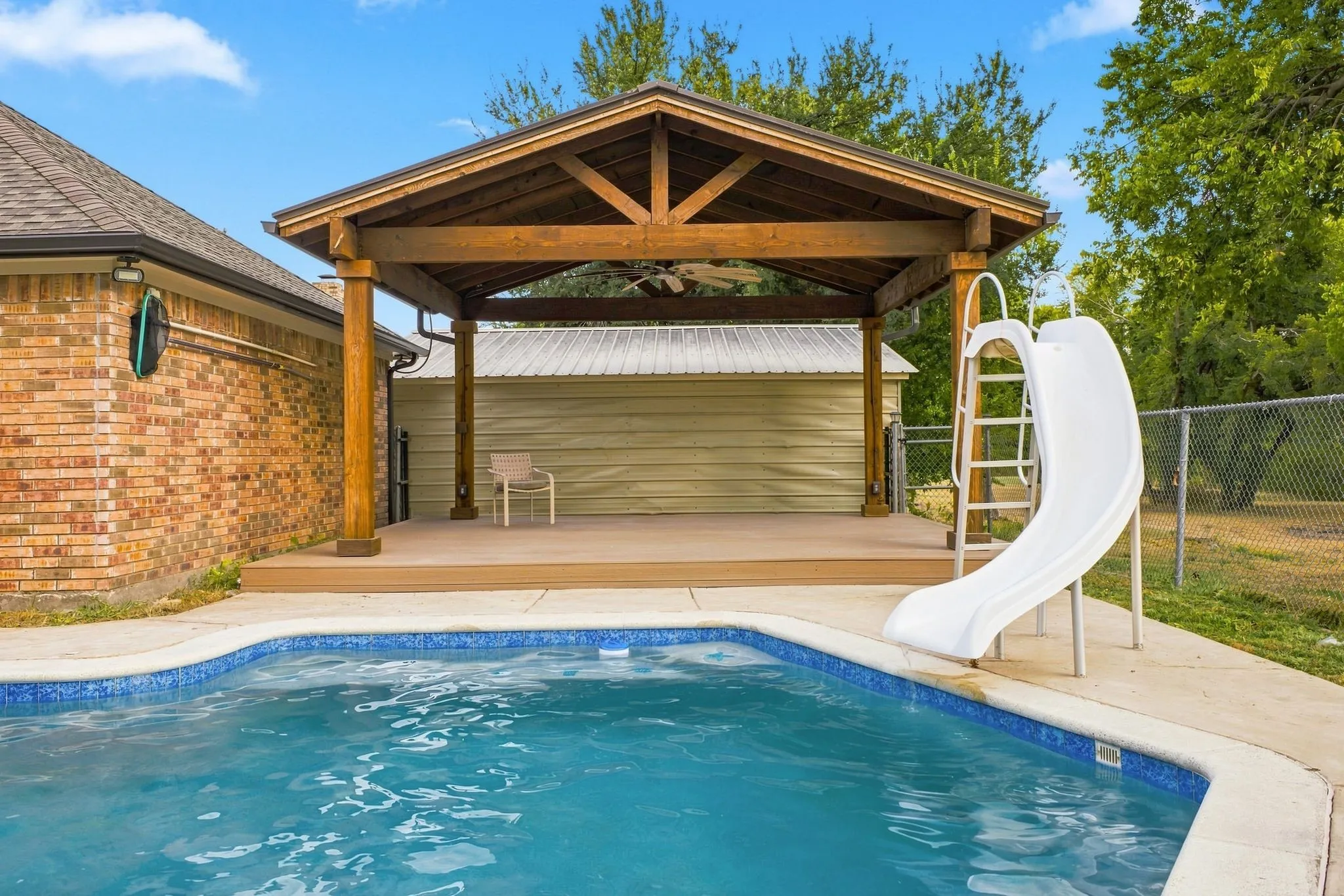 Outdoor swimming pool with a concrete surround, featuring a wooden pergola-covered deck with exposed beams and a ceiling fan