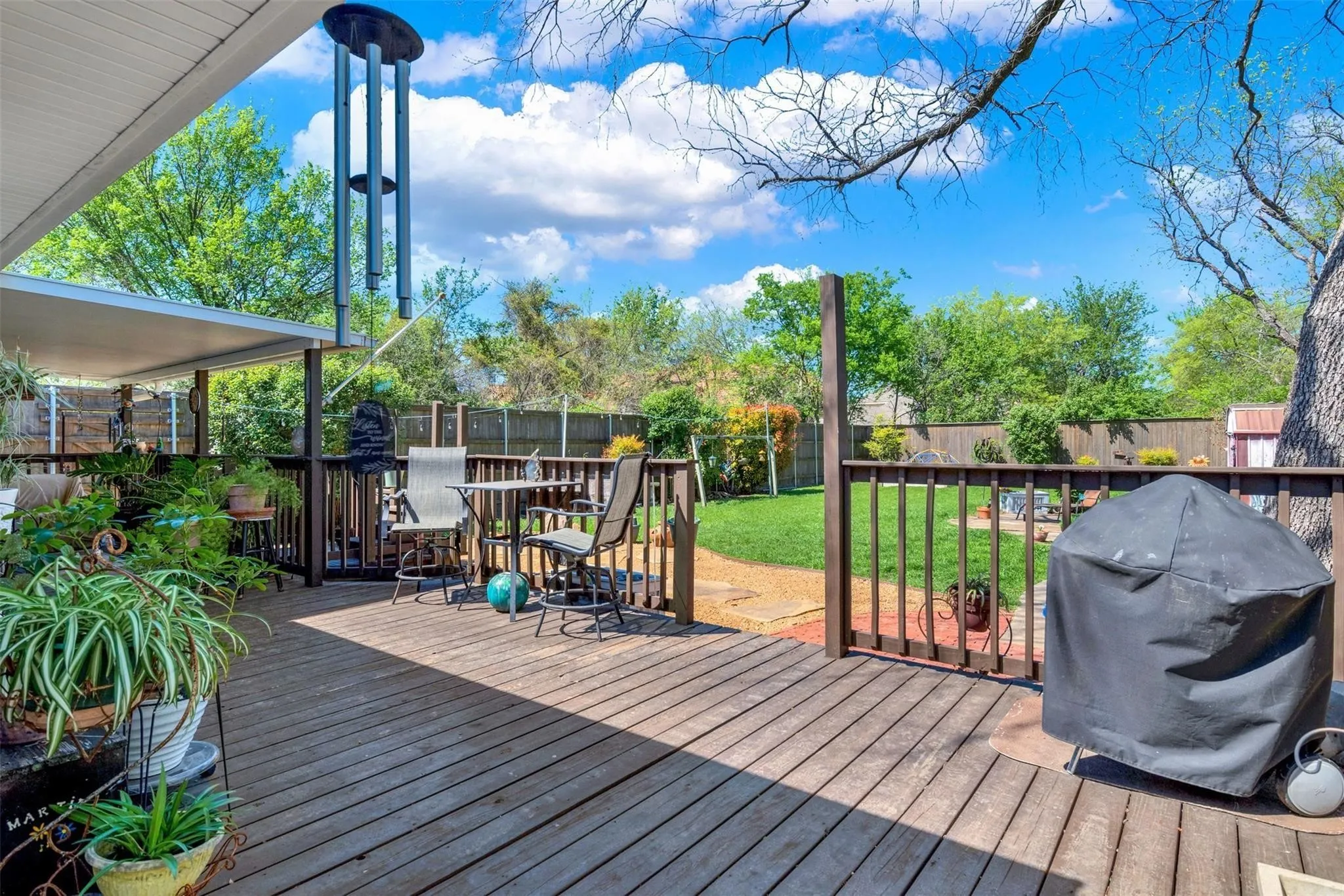 Expansive wood deck with a covered patio area, dark wood railing, stone paver pathway, and mature trees