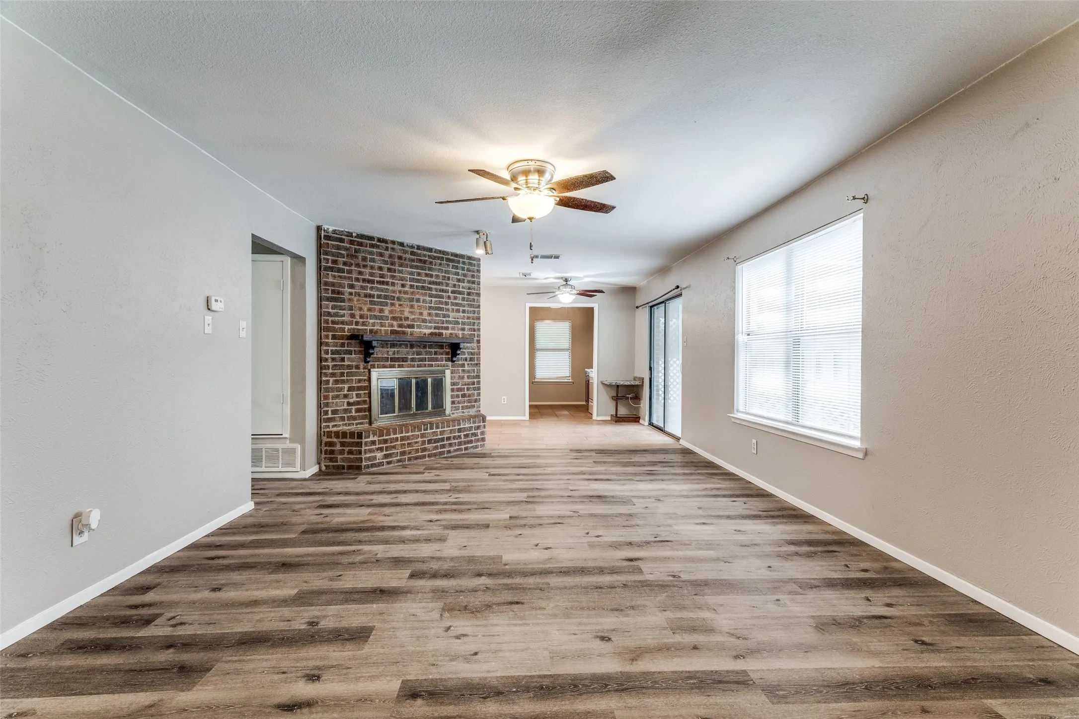 Wood-finish flooring, brick fireplace with mantel, ceiling fans, and large window with blinds