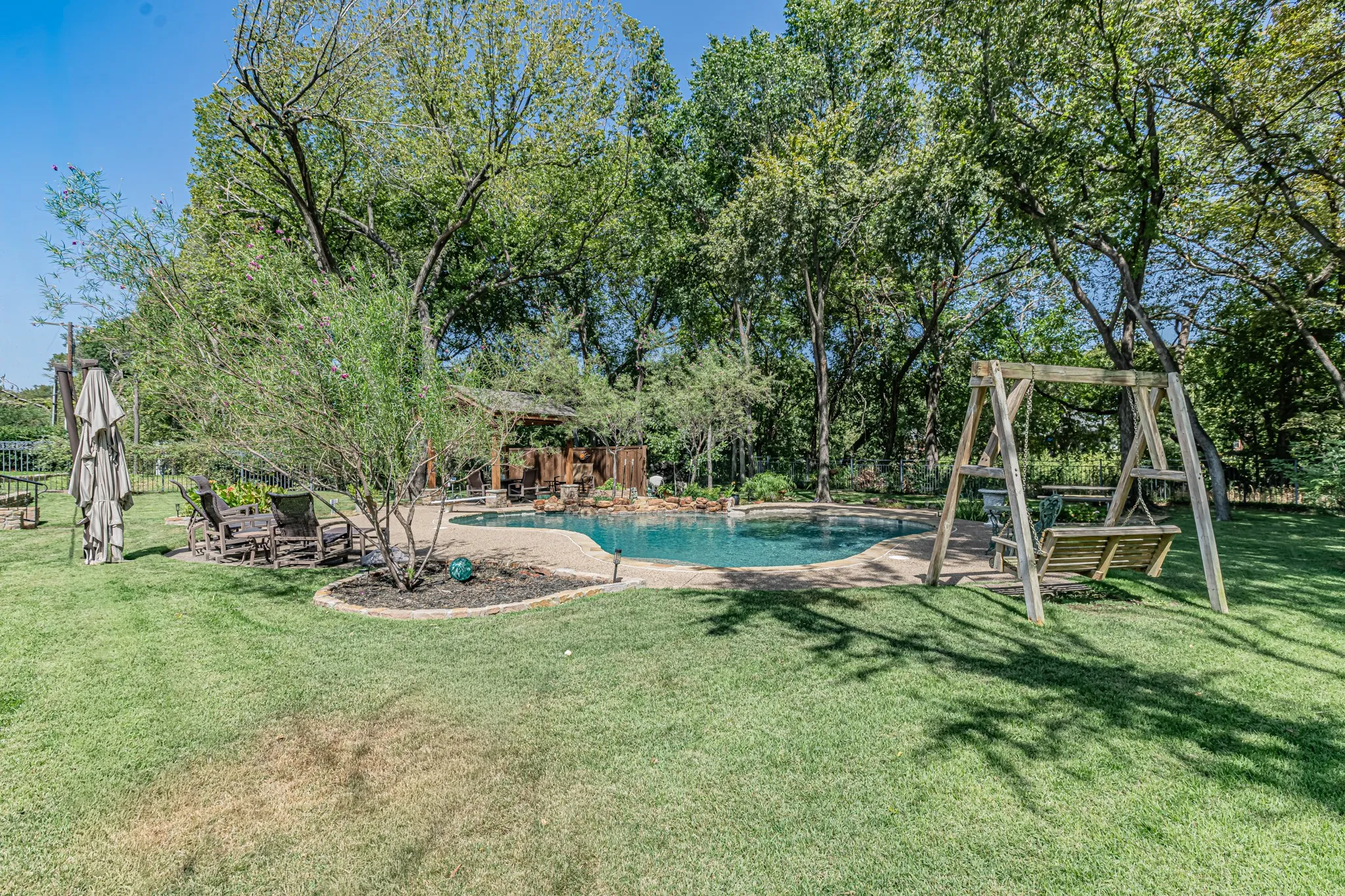 Spacious backyard featuring a freeform swimming pool, extensive stone hardscaping, mature trees, and a built-in gazebo