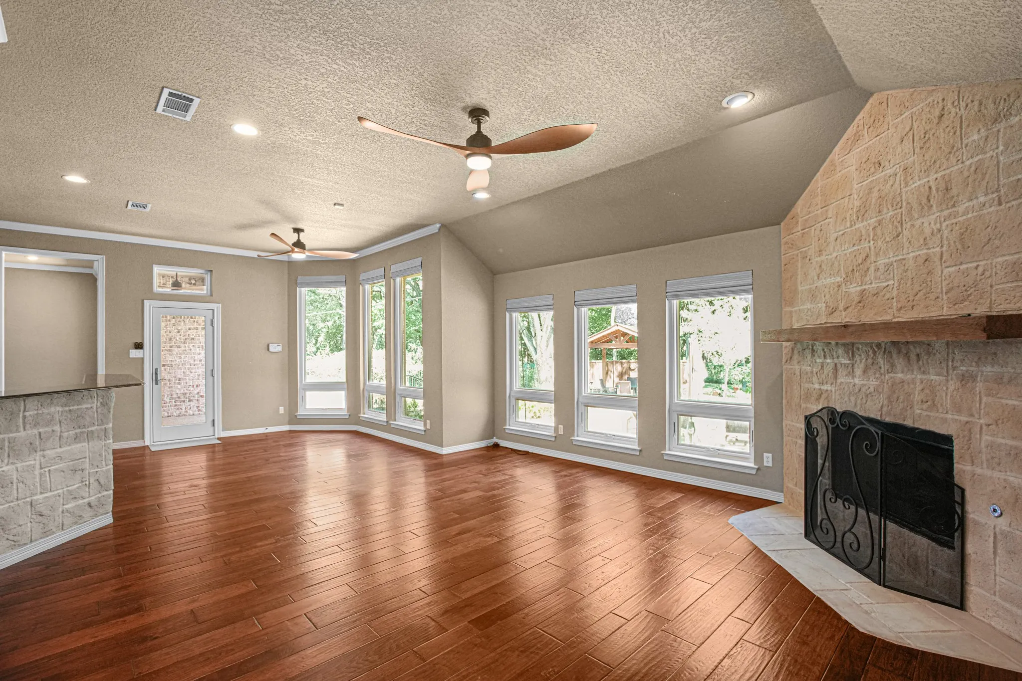 Vaulted ceiling with recessed lighting, wood-finish flooring, stone-clad fireplace with mantel, multiple large windows, and an accent stone-clad bar