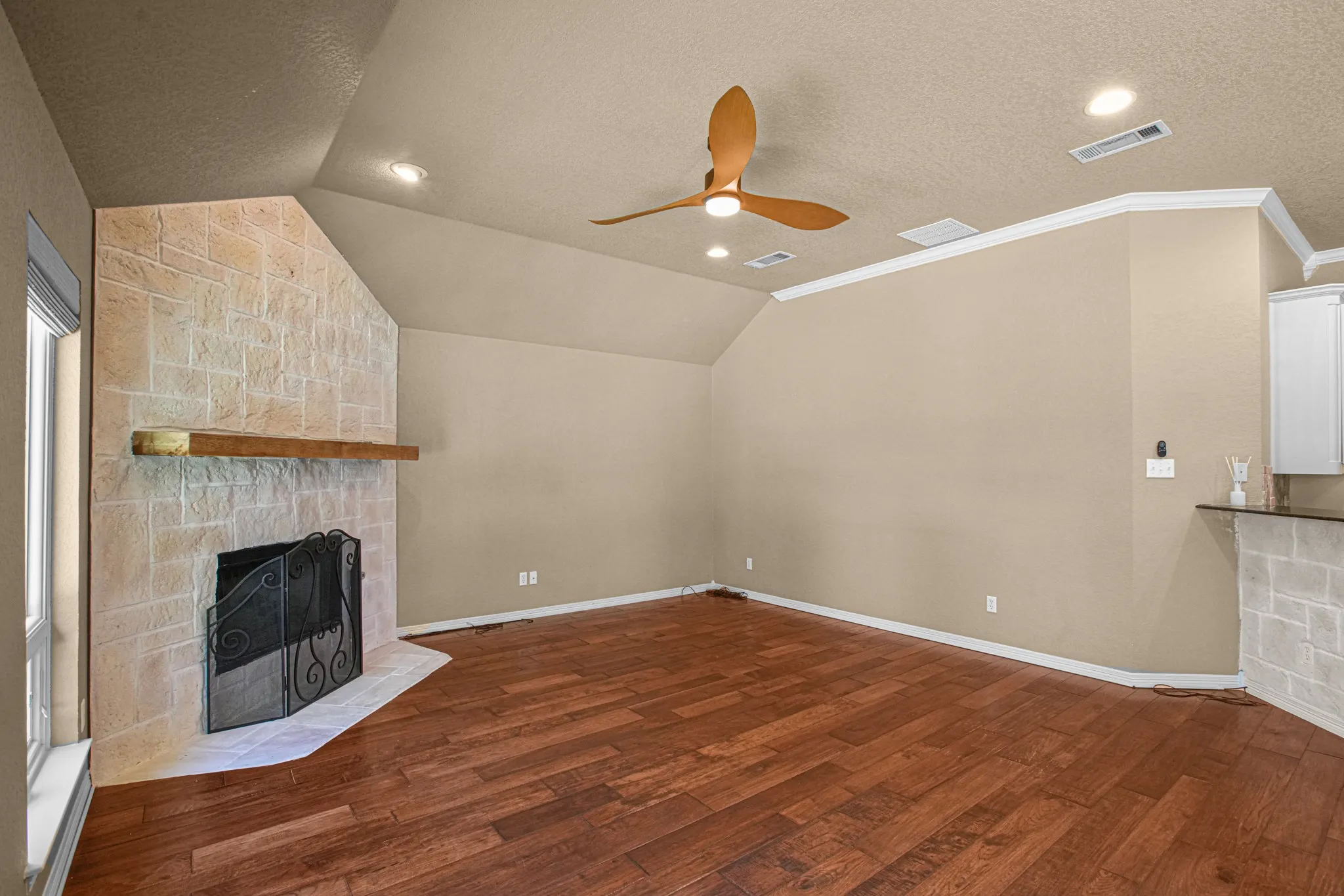 Vaulted ceiling with recessed lighting and a ceiling fan, a stone-clad fireplace with a wood mantle, wood-finish flooring, and crown molding