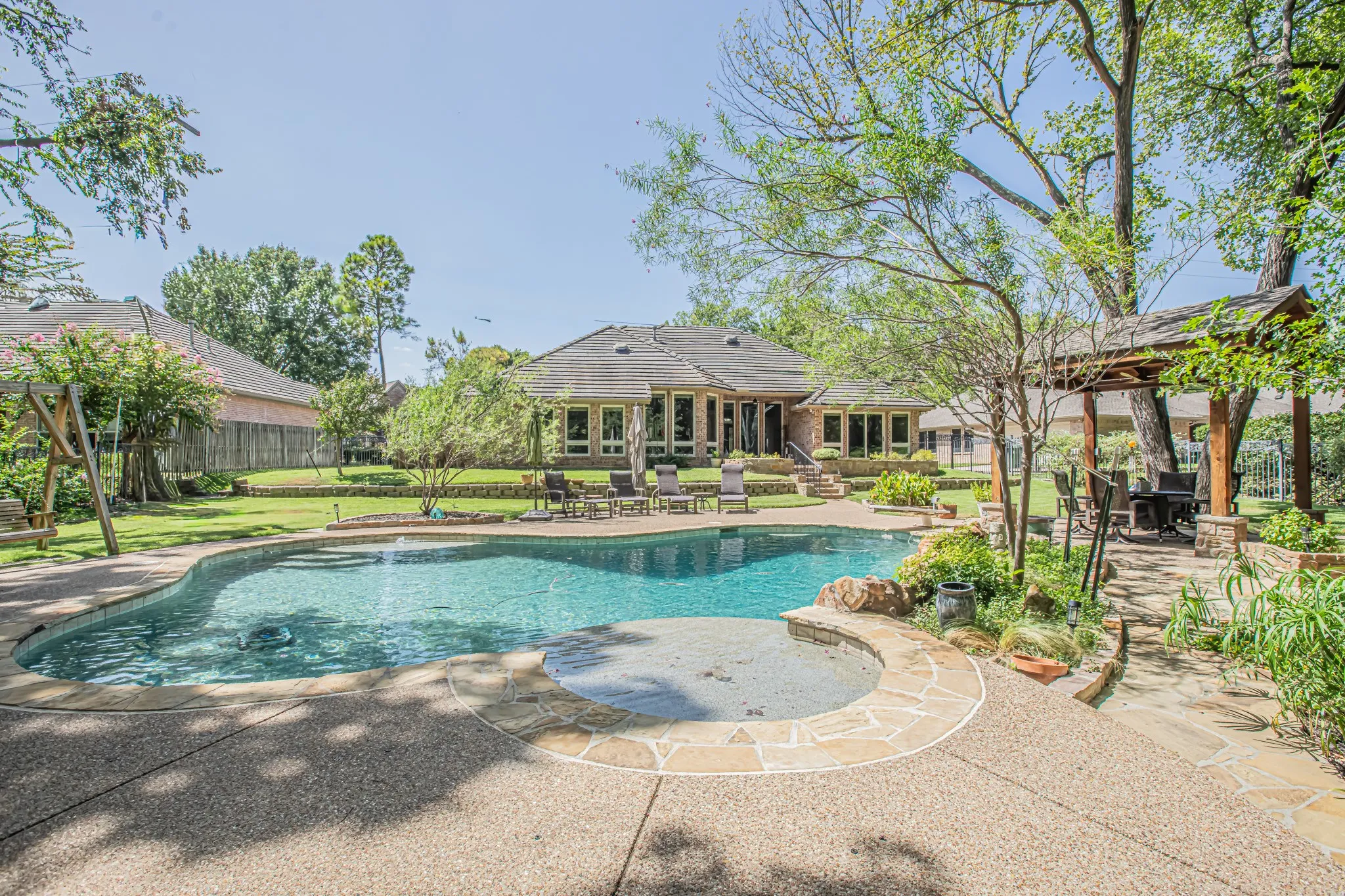 Expansive backyard featuring a freeform swimming pool with integrated spa, stone-edged landscaping, exposed aggregate decking, and a covered gazebo