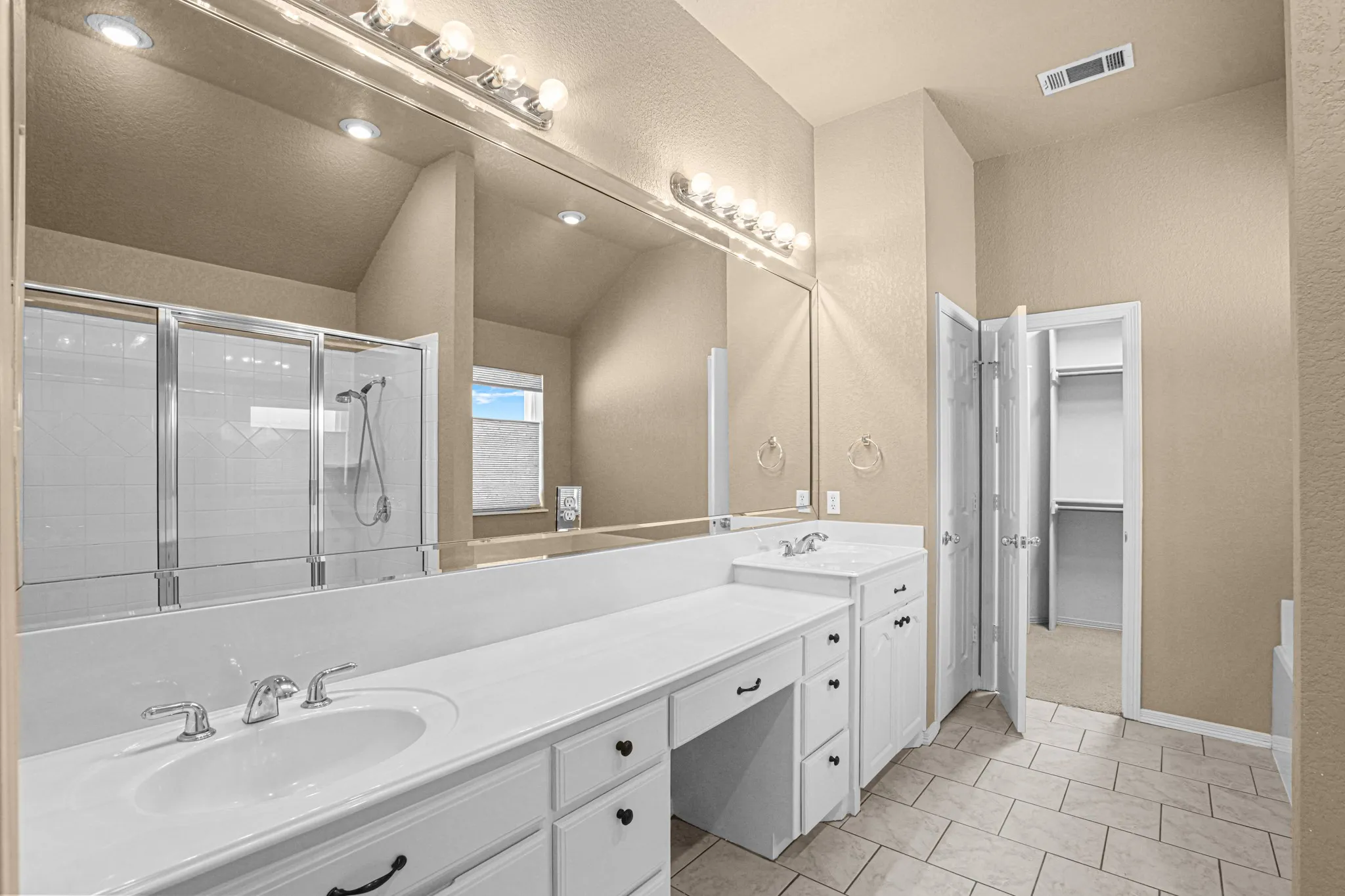 Dual vanity with white cabinetry and integrated sinks, large mirrored wall with overhead lighting, tiled flooring, and a glass-enclosed shower