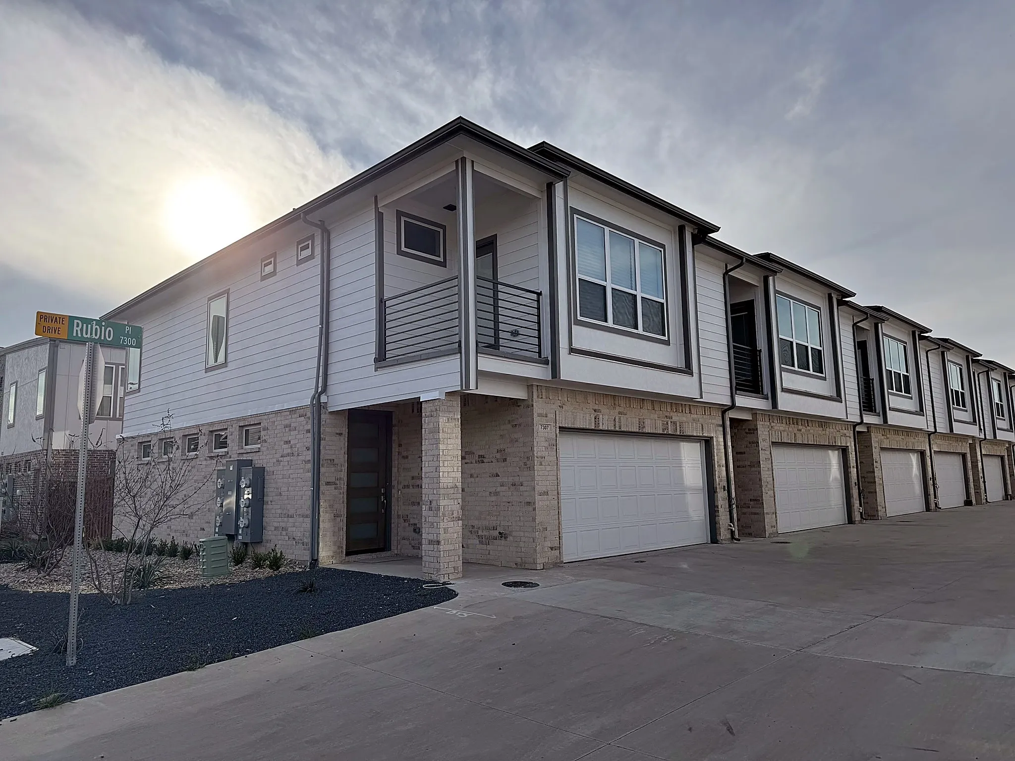 Modern townhouse with light-toned siding and brick veneer, featuring an integrated garage, large windows with dark trim, and a second-story balcony with metal railing