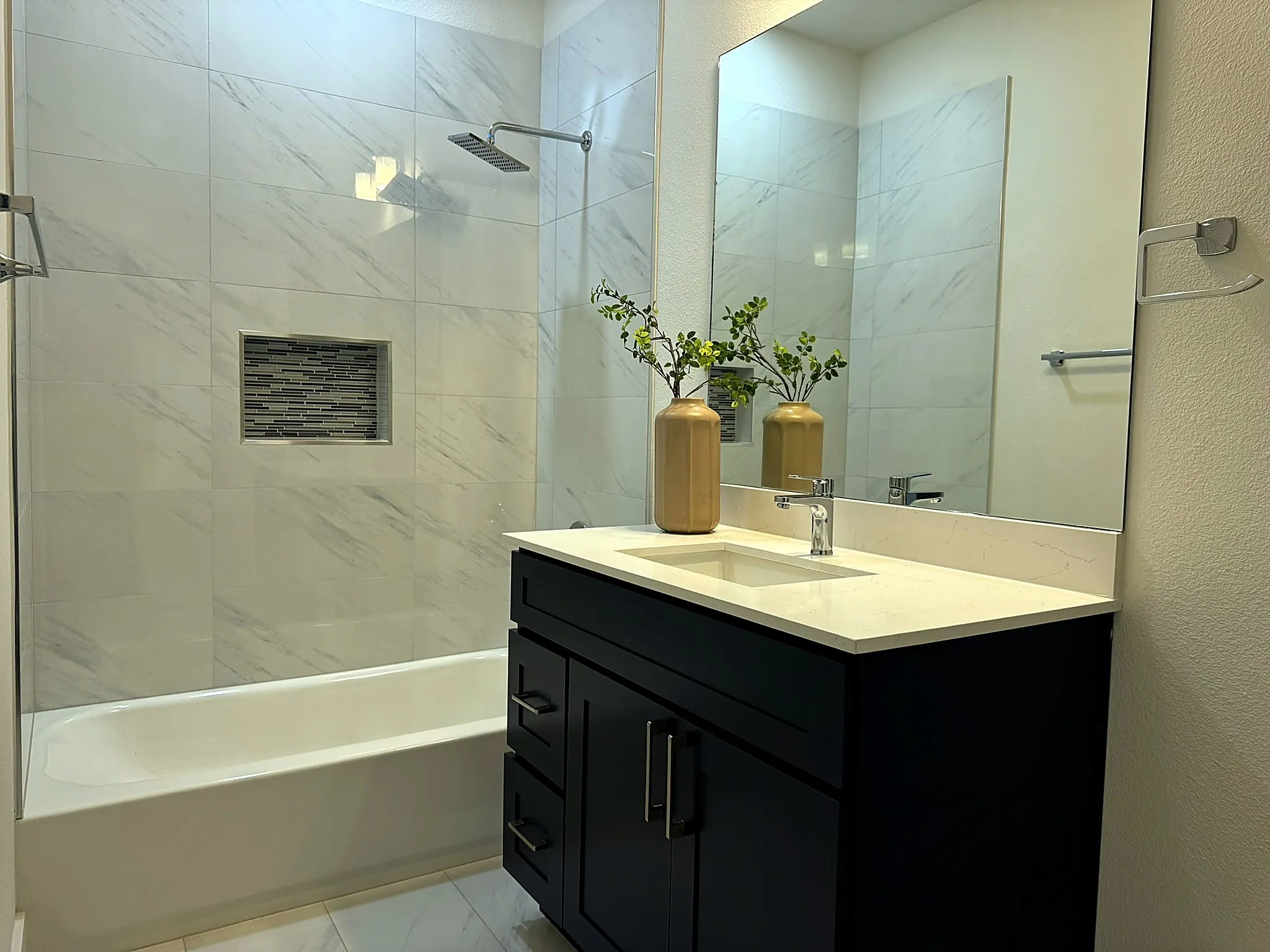 Bathroom featuring a vanity with an integrated sink, a large mirror, a chrome faucet, and a shower/tub combination with white tile surround and an inset mosaic tile niche