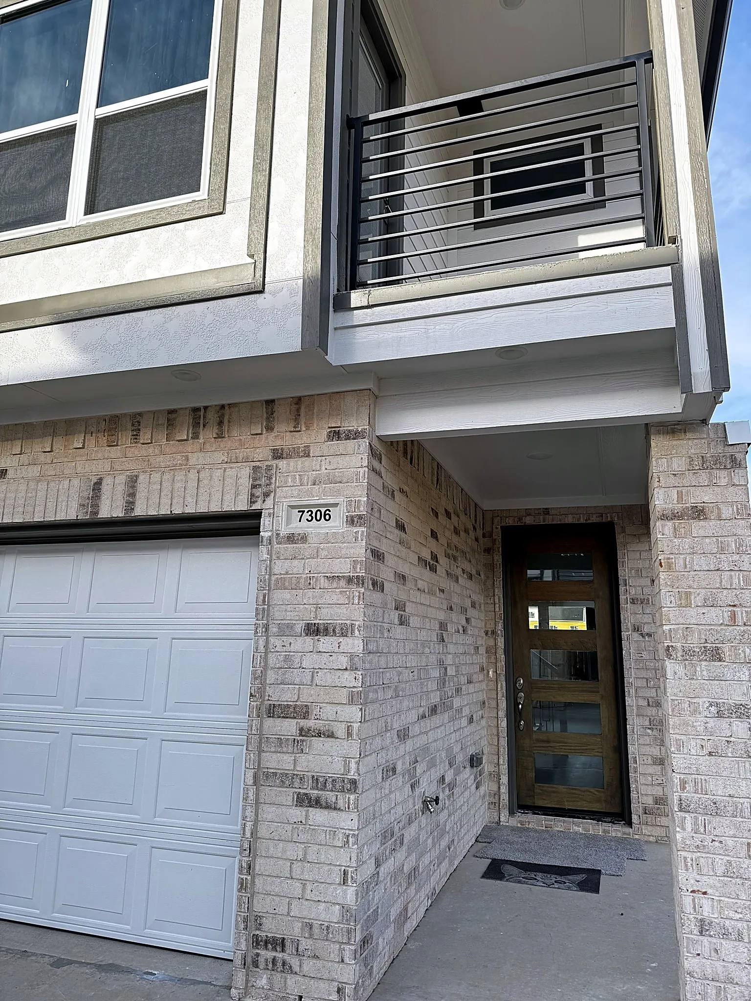 Light-colored brick exterior with a modern wood-panel entry door, white panel garage door, and an upper-level balcony featuring horizontal metal railings