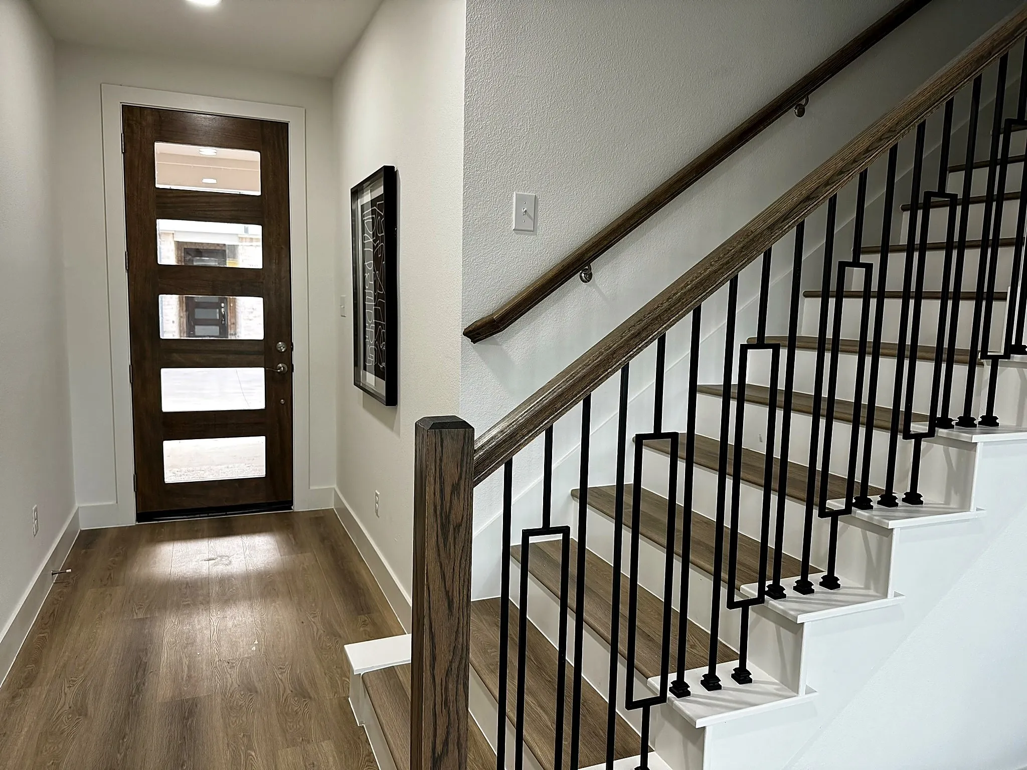 Wood-finish flooring, contemporary wood front door with glass panels, recessed lighting, and a staircase featuring a wood handrail with decorative metal balusters