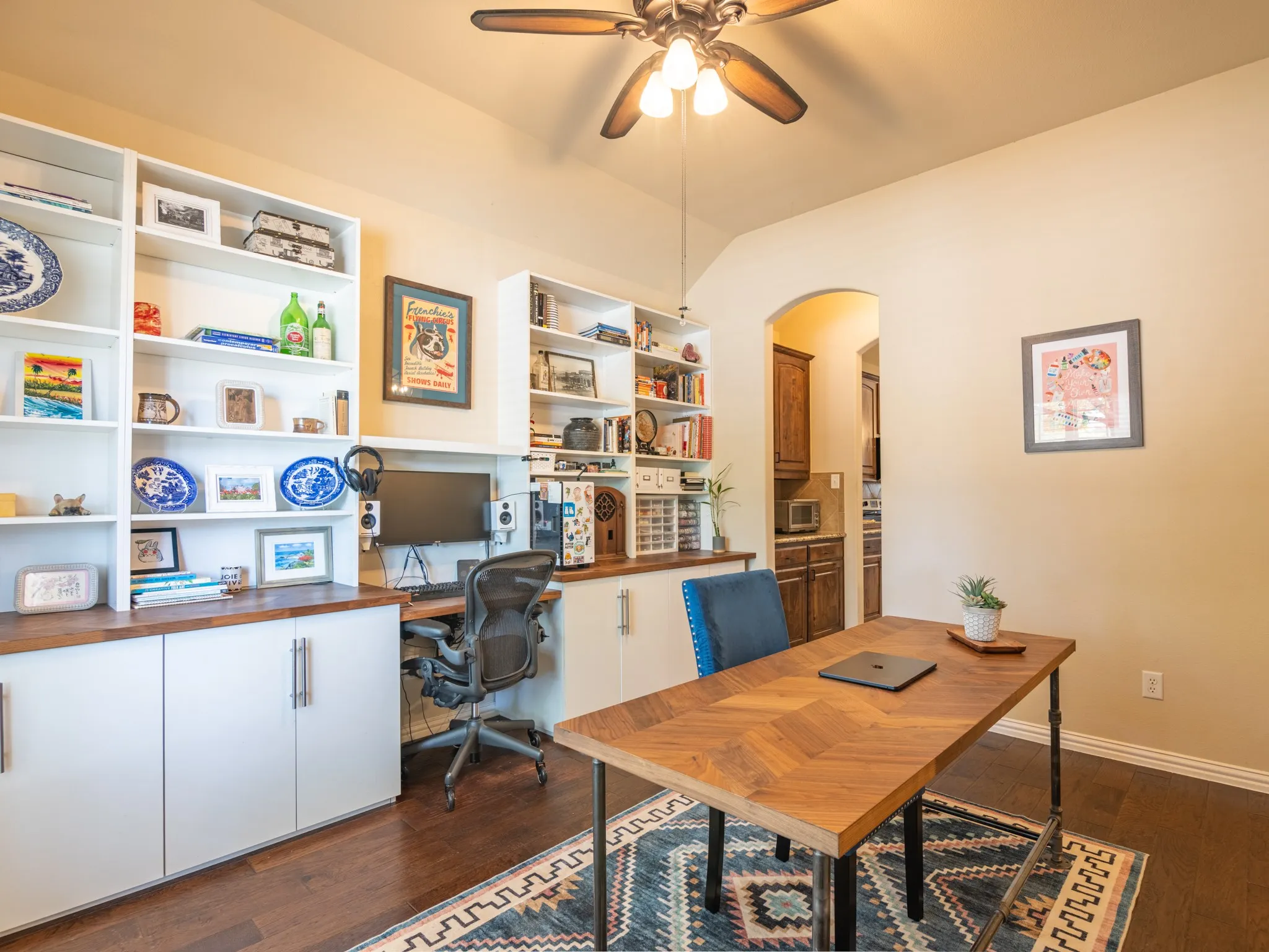 Hardwood-finish flooring, built-in shelving, recessed workspace with cabinet storage, overhead ceiling fan, and an arched doorway