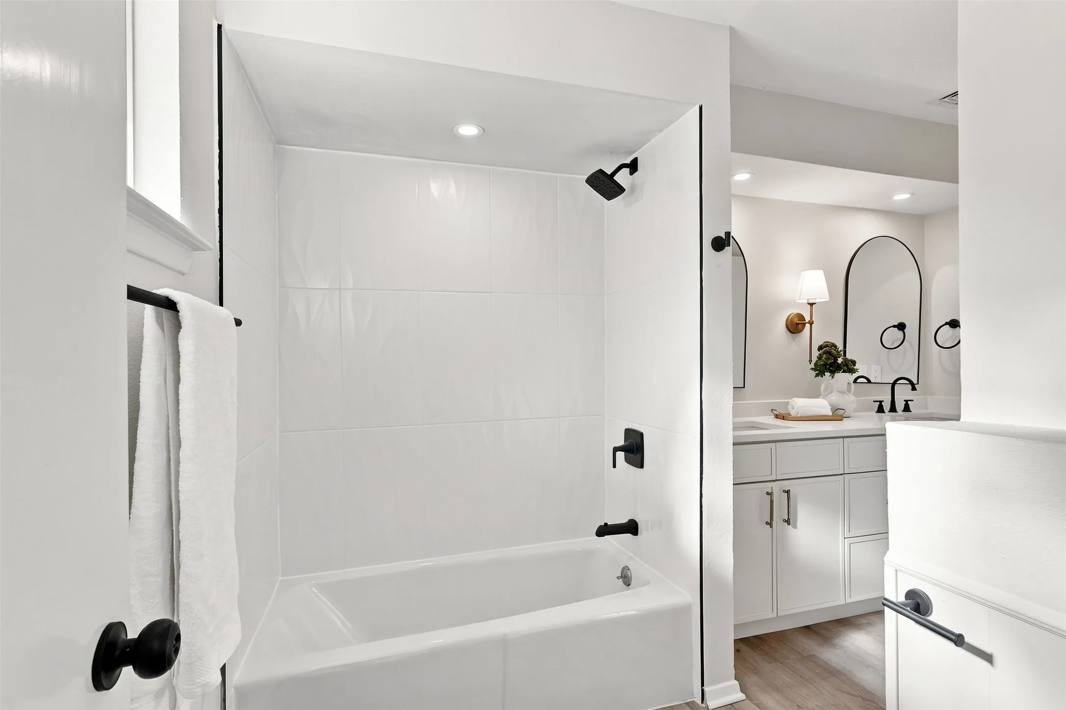Bathtub with white tile surround and matte black fixtures, recessed lighting, and wood-finish flooring