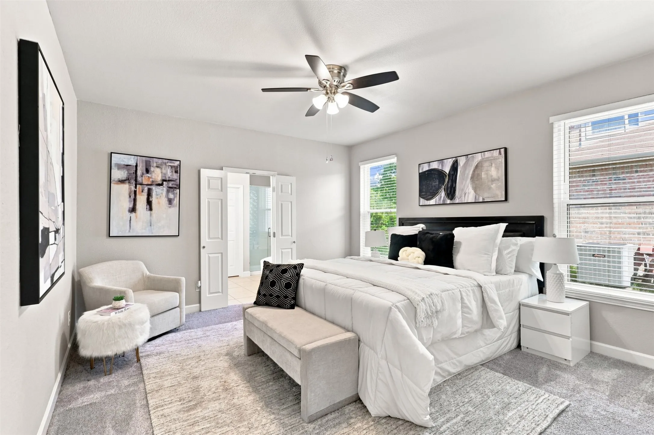Spacious primary bedroom featuring light walls, carpeted flooring, dual windows with blinds, a ceiling fan with integrated lighting, and white paneled double doors leading to the en-suite bathroom.
