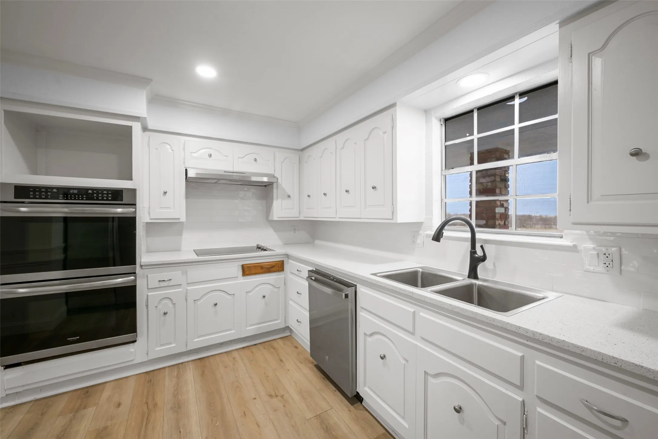 Kitchen featuring white cabinetry, light-toned countertops, wood-finish flooring, a double wall oven, and a stainless steel double basin sink with a black faucet