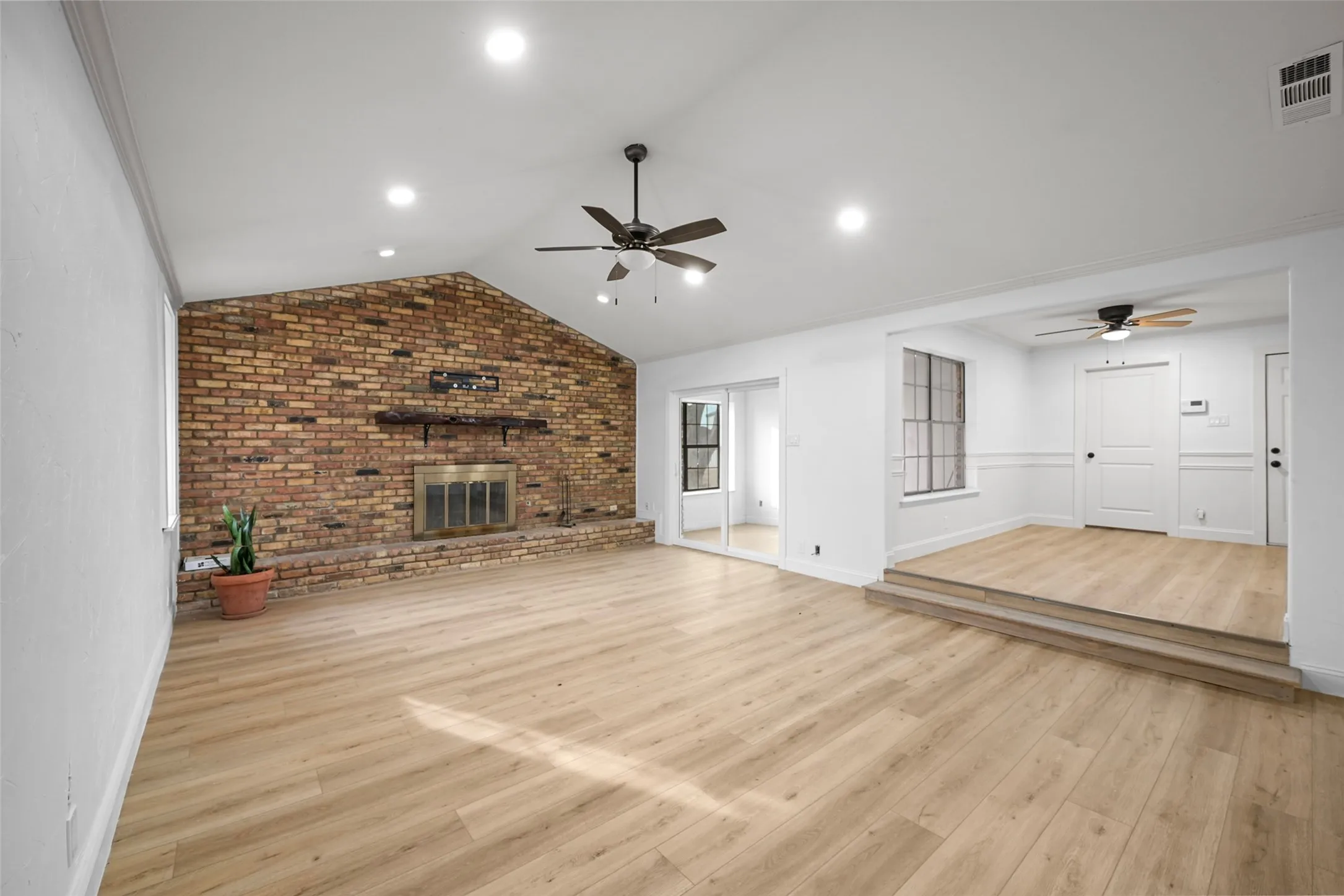Vaulted ceiling with recessed lighting, brick-faced fireplace with hearth, wood-finish flooring, two ceiling fans, and an elevated room