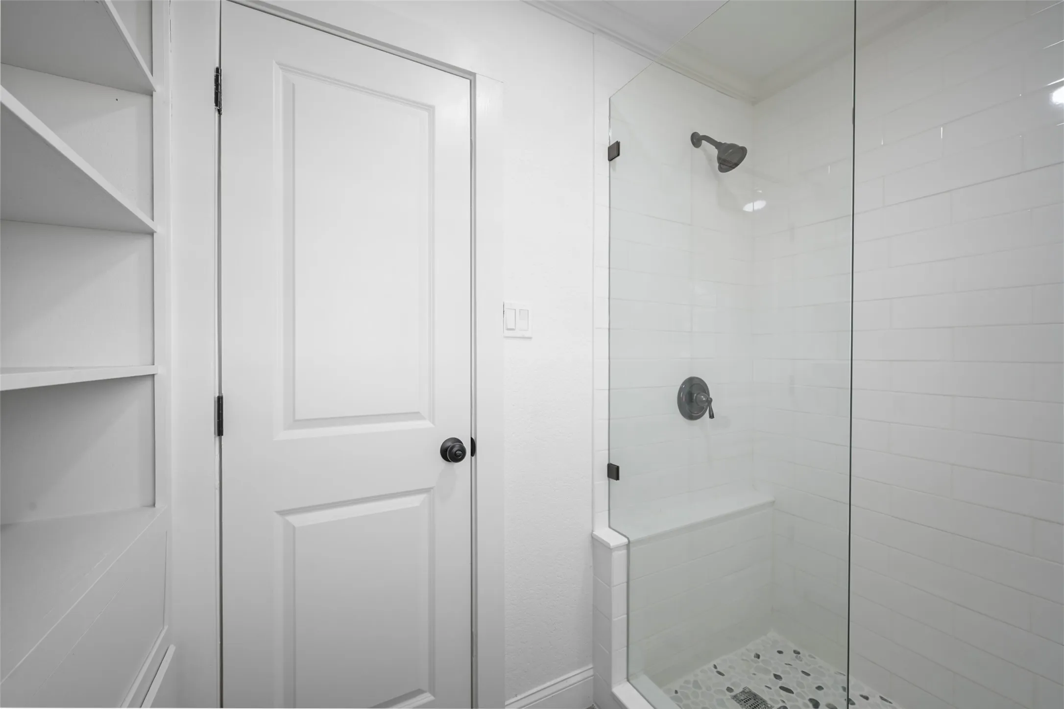 Built-in shelving, white paneled door, glass-enclosed shower with subway tile surround, integrated shower bench, and pebble-style shower pan