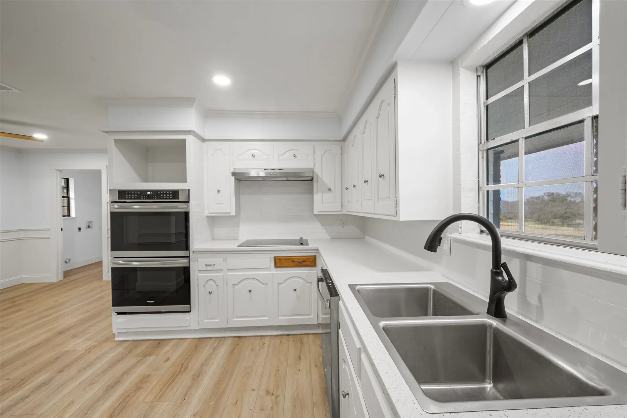 Kitchen with wood-finish flooring, white cabinetry, integrated double ovens, and a dual basin sink with a matte black faucet