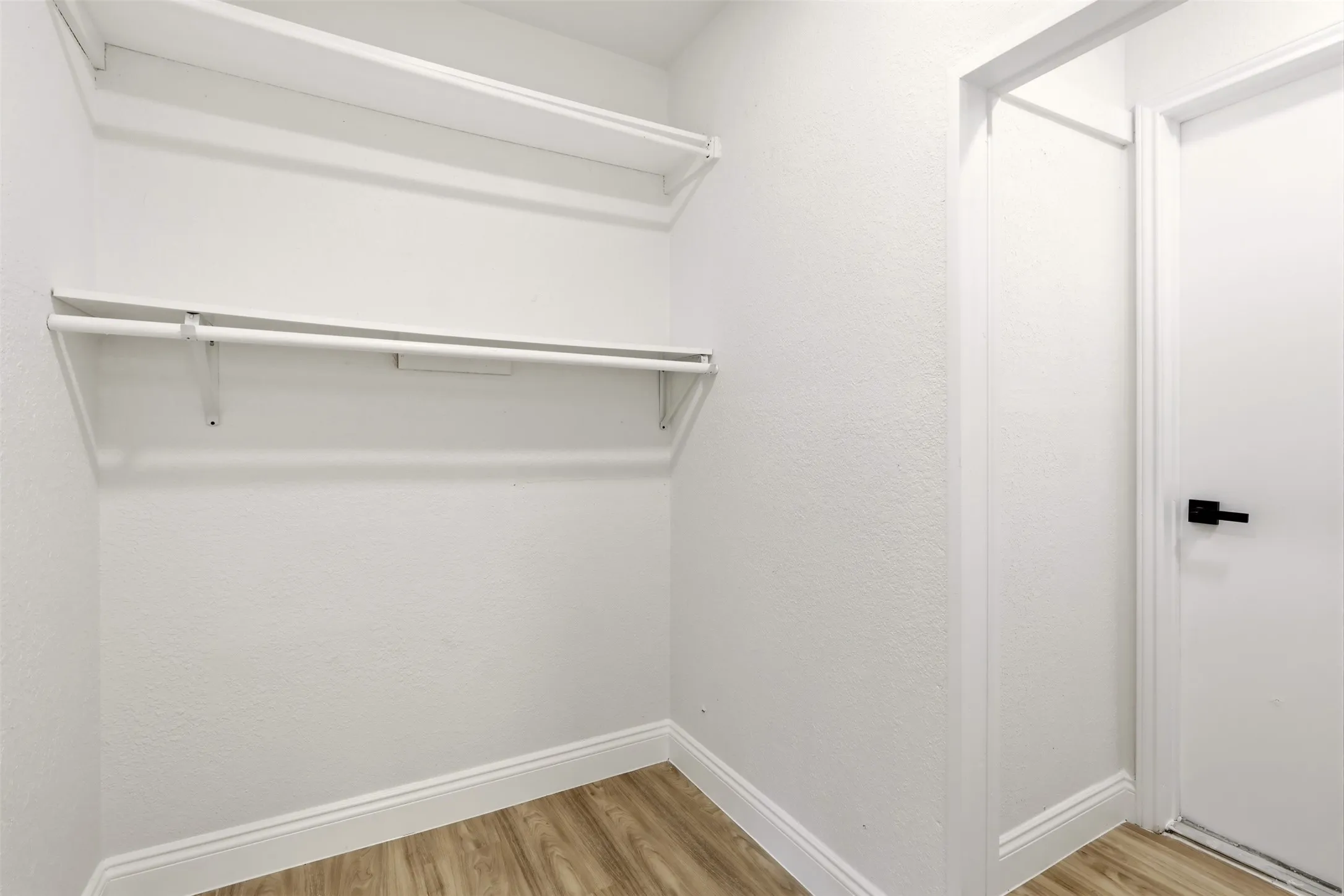 Wood-finish flooring with white baseboards, white walls, and a white door with a black handle