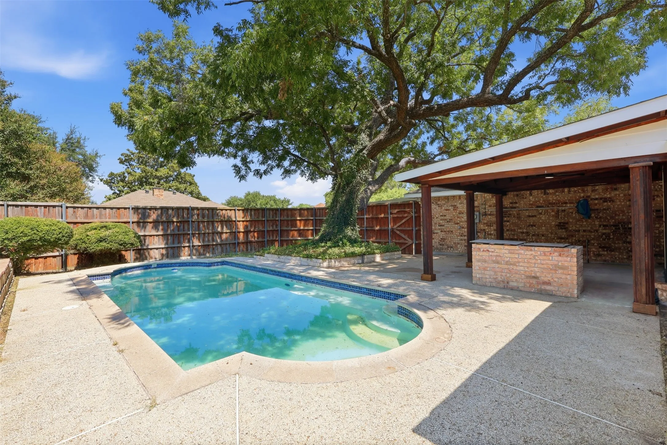 Resort-style backyard oasis featuring a sparkling free-form swimming pool with shallow lounging areas, expansive concrete decking, mature specimen tree providing natural shade, brick and wood-finish covered patio structure, and mature landscaping with brick perimeter fencing