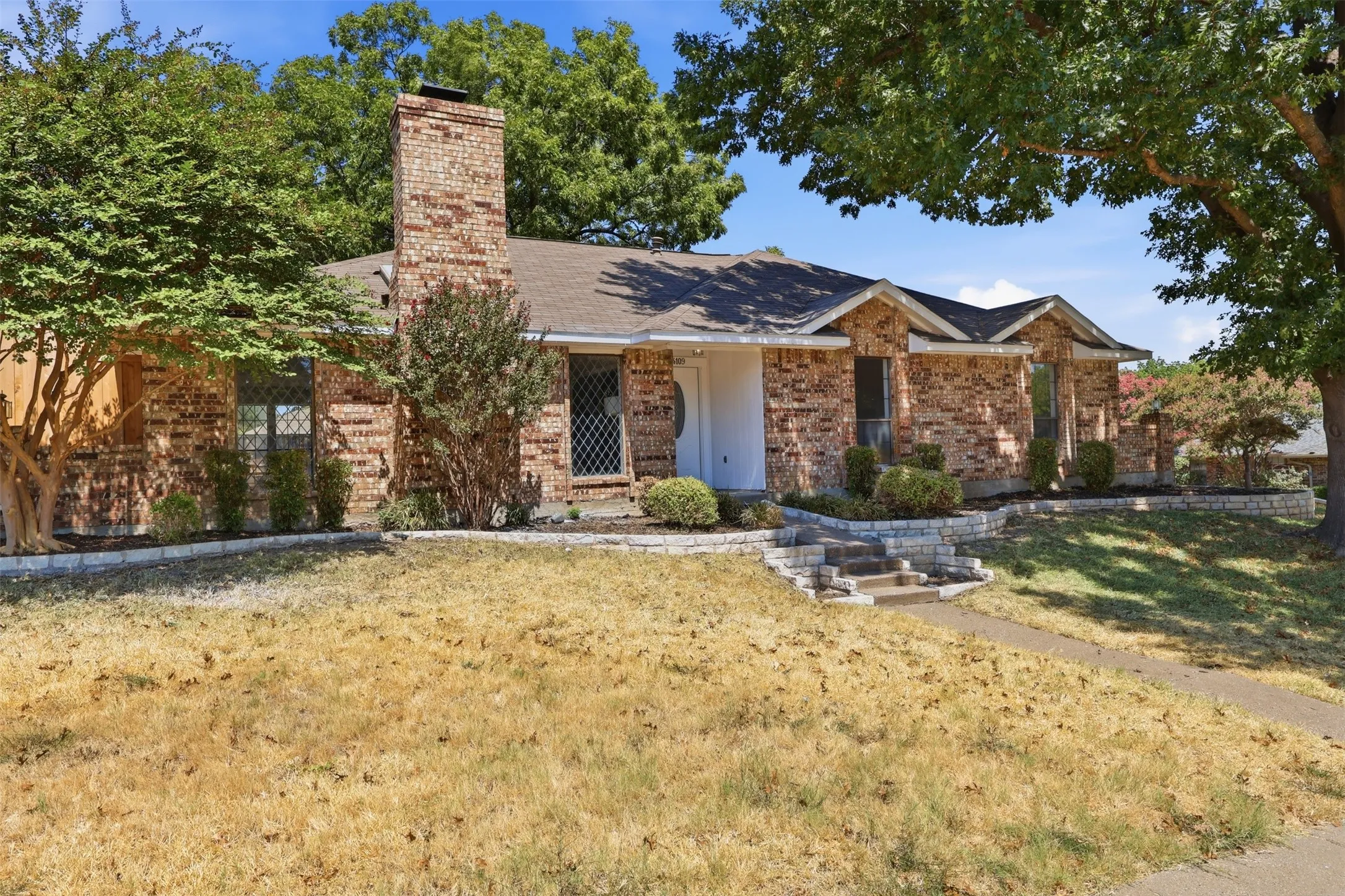 Brick facade with a prominent chimney, stone-edged landscaping, and mature trees