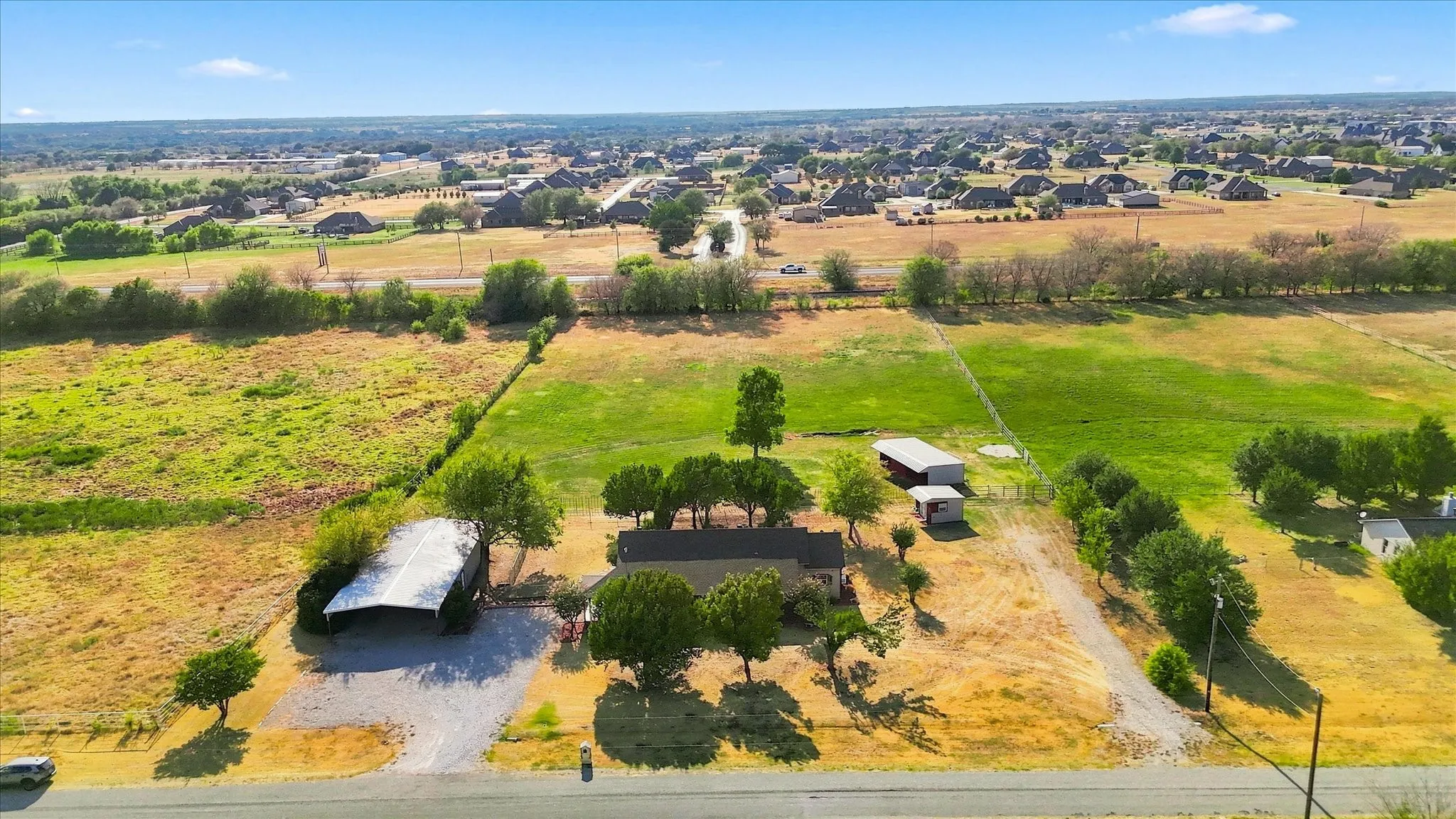 Expansive property featuring a residence with a dark roof, surrounded by mature trees, multiple outbuildings, and a large open field